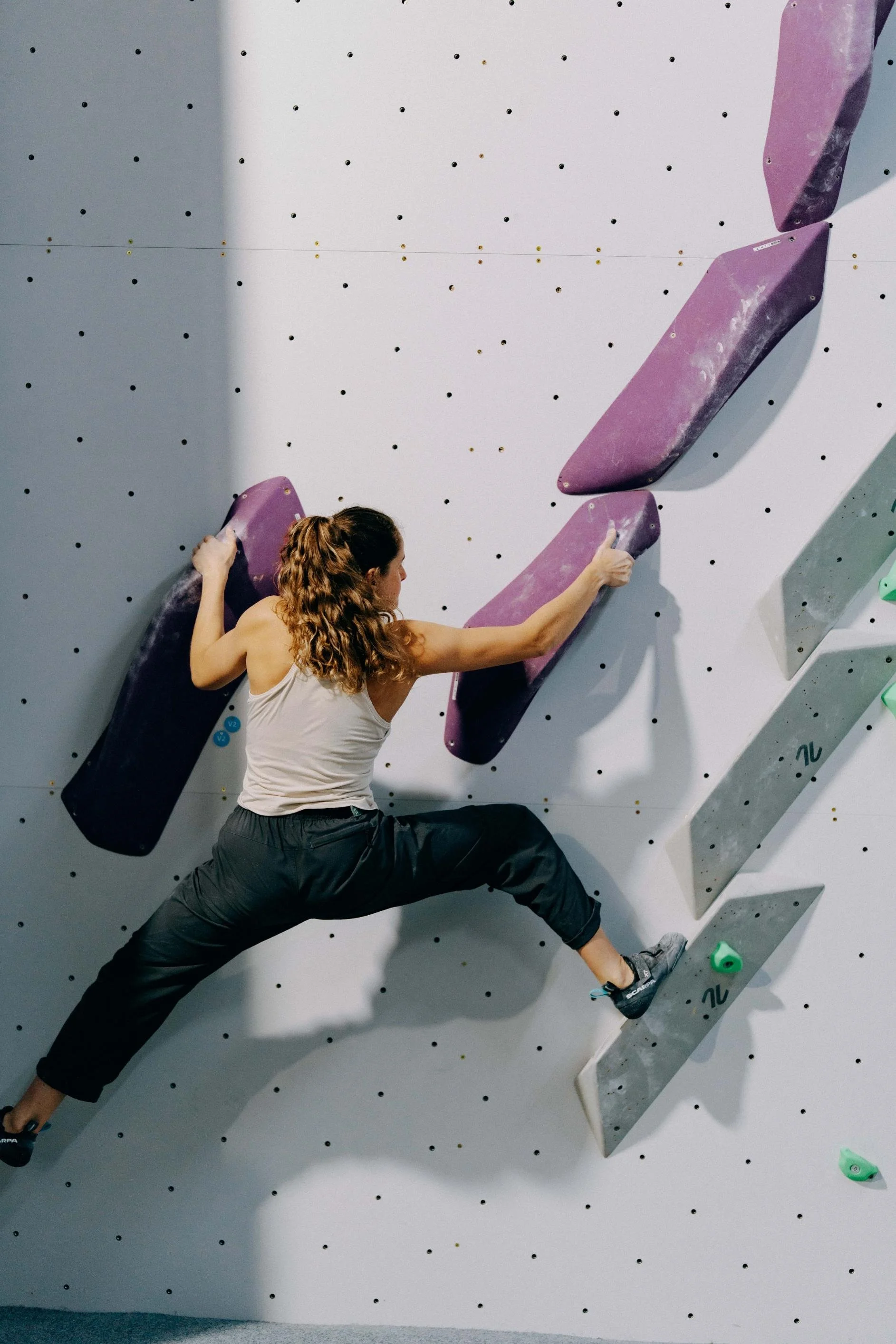 A woman with long curly hair wearing a white tank top, black pants, and climbing shoes is practicing indoor rock climbing on a white wall with purple holds.