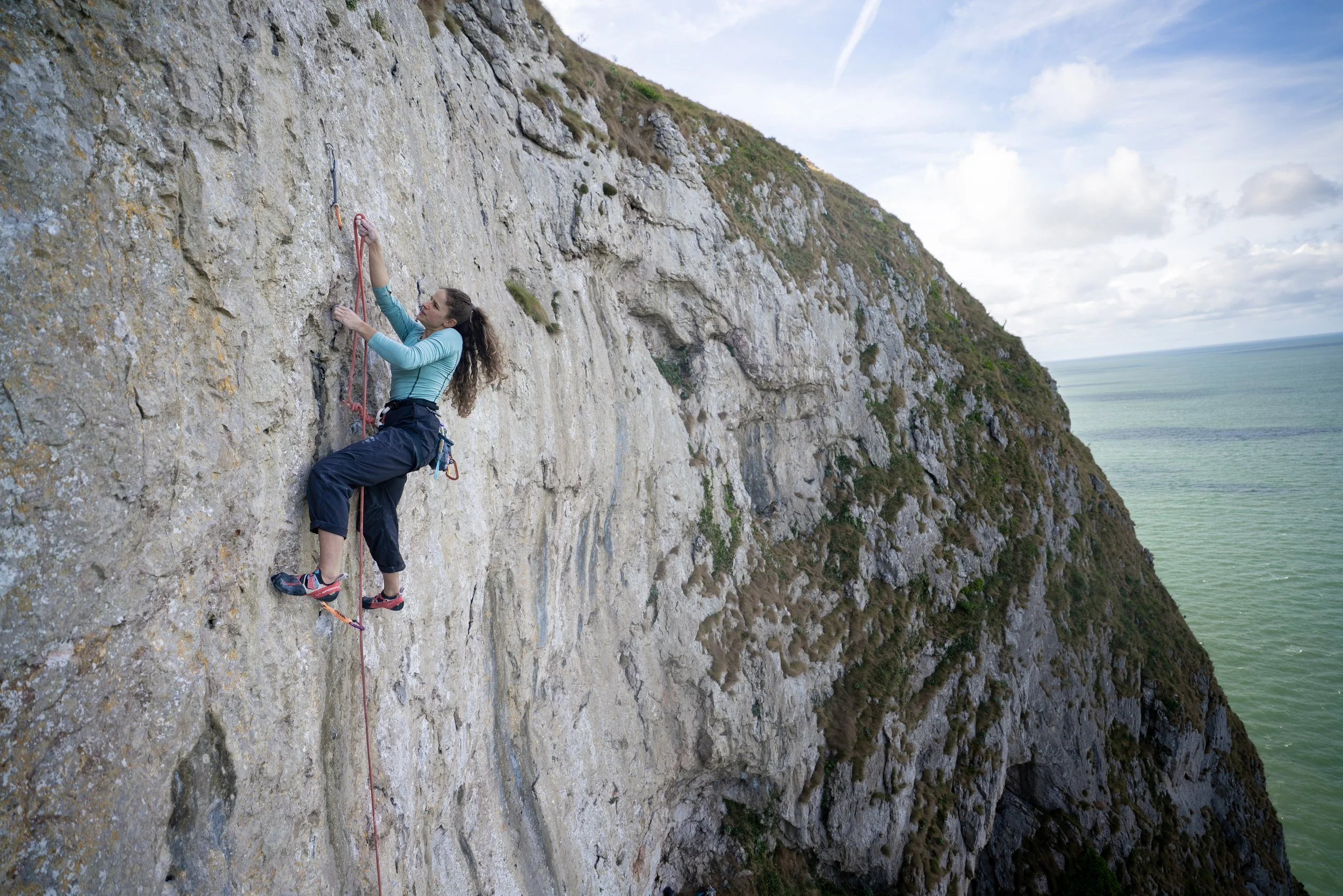 A woman rock climbing on a steep cliff face overlooking the ocean, with a cloudy sky above.