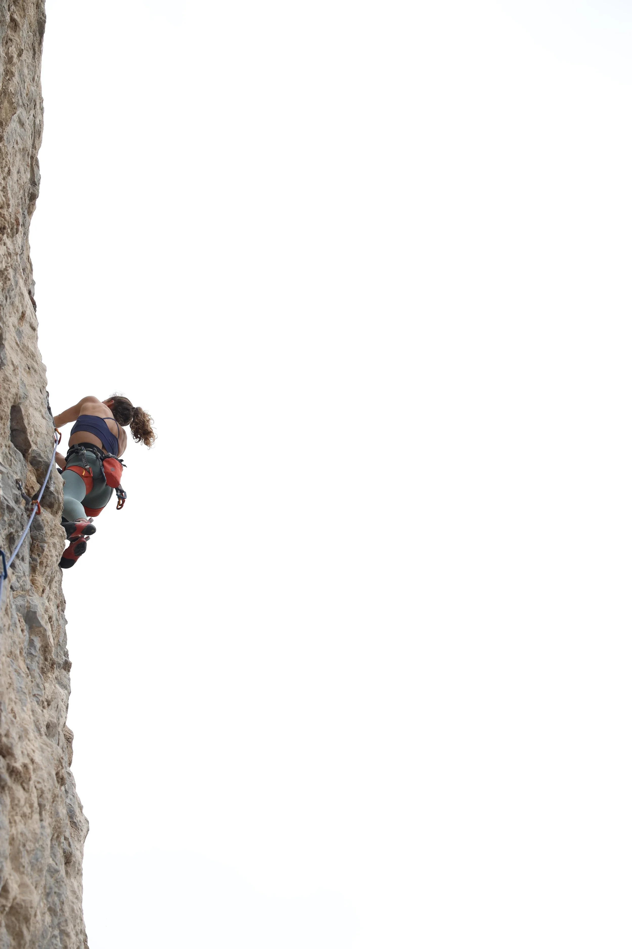 A person rock climbing on an outdoor cliff face, wearing a harness and climbing shoes, with a cloudy sky in the background.