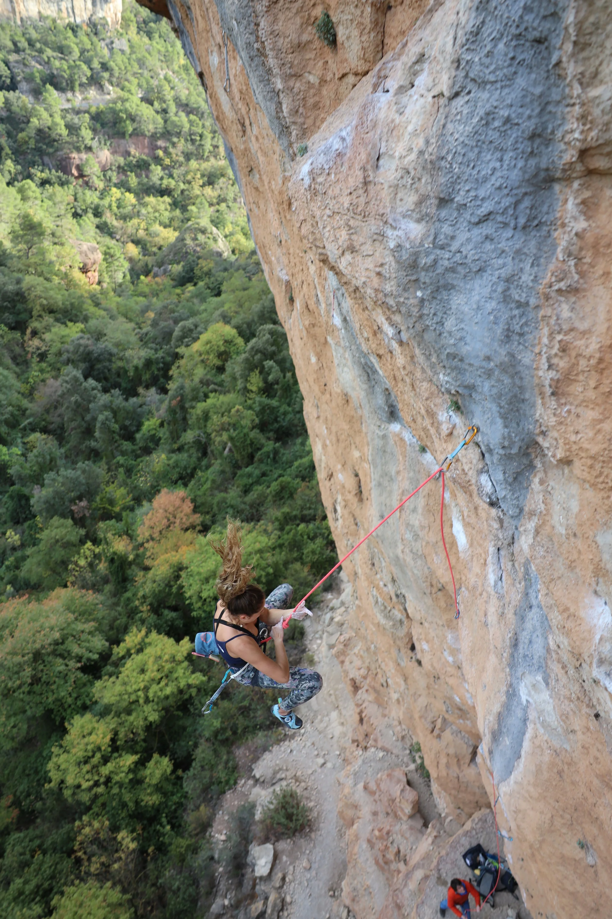 A woman rock climbing on a steep outdoor cliff face with trees and a valley in the background.