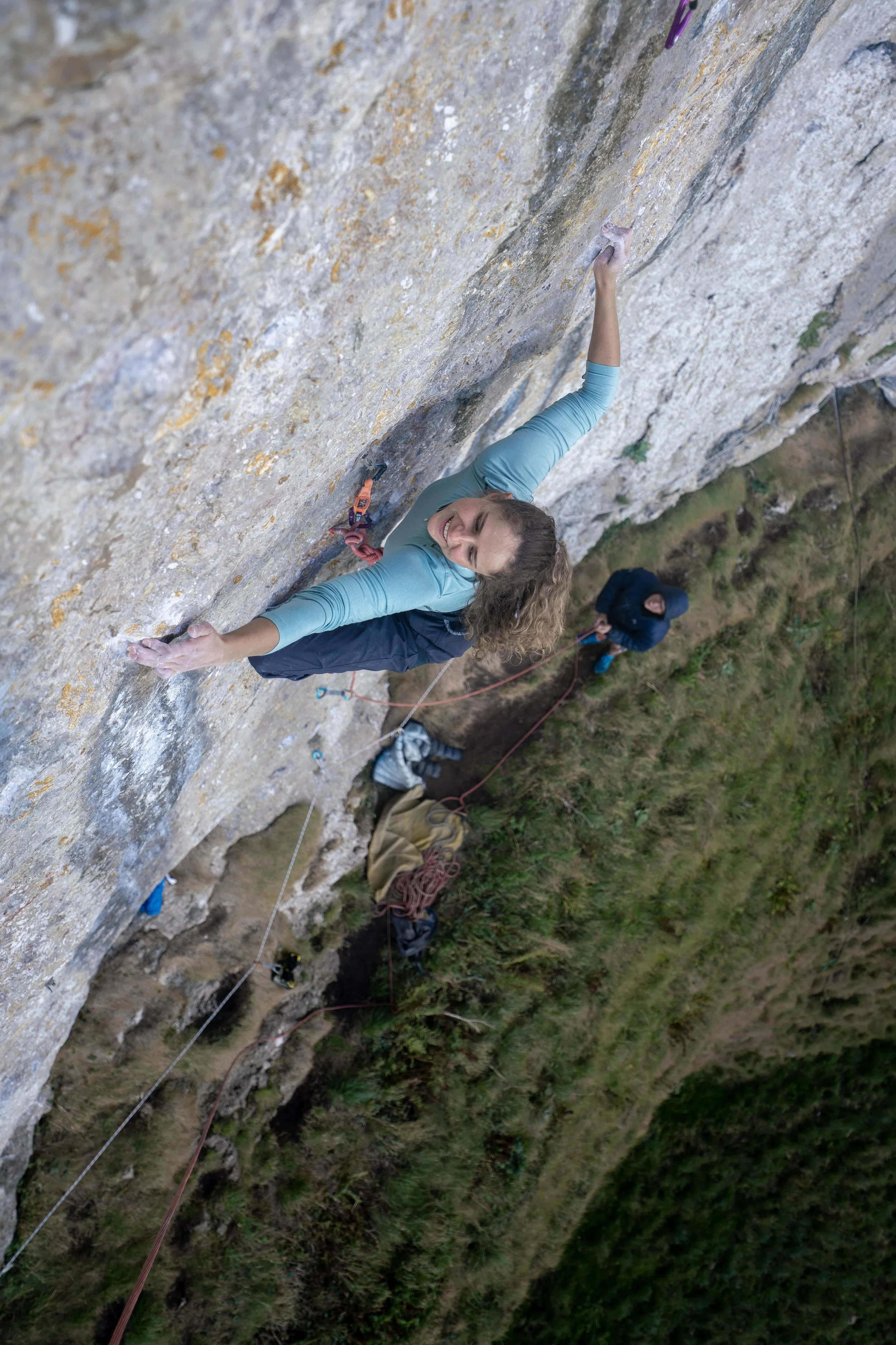 A woman is smiling as she climbs a steep outdoor rock face with climbing gear, with another person belaying her from below on a grassy and wooded terrain.