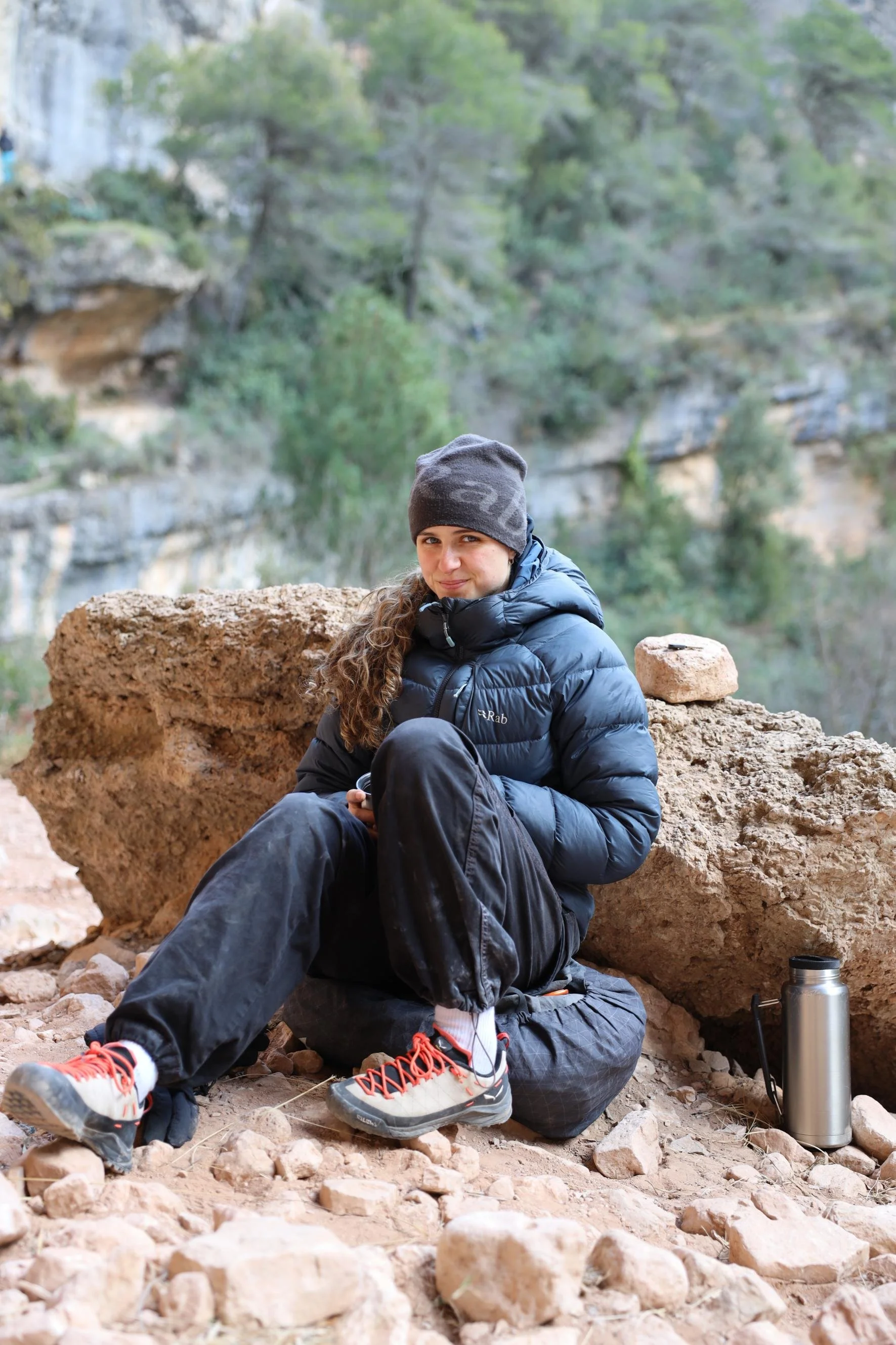 A woman sitting on rocky terrain in an outdoor setting, wearing a gray beanie, black puffer jacket, black pants, and hiking shoes, with a water bottle and a thermos nearby. She is surrounded by rocks and greenery in the background.