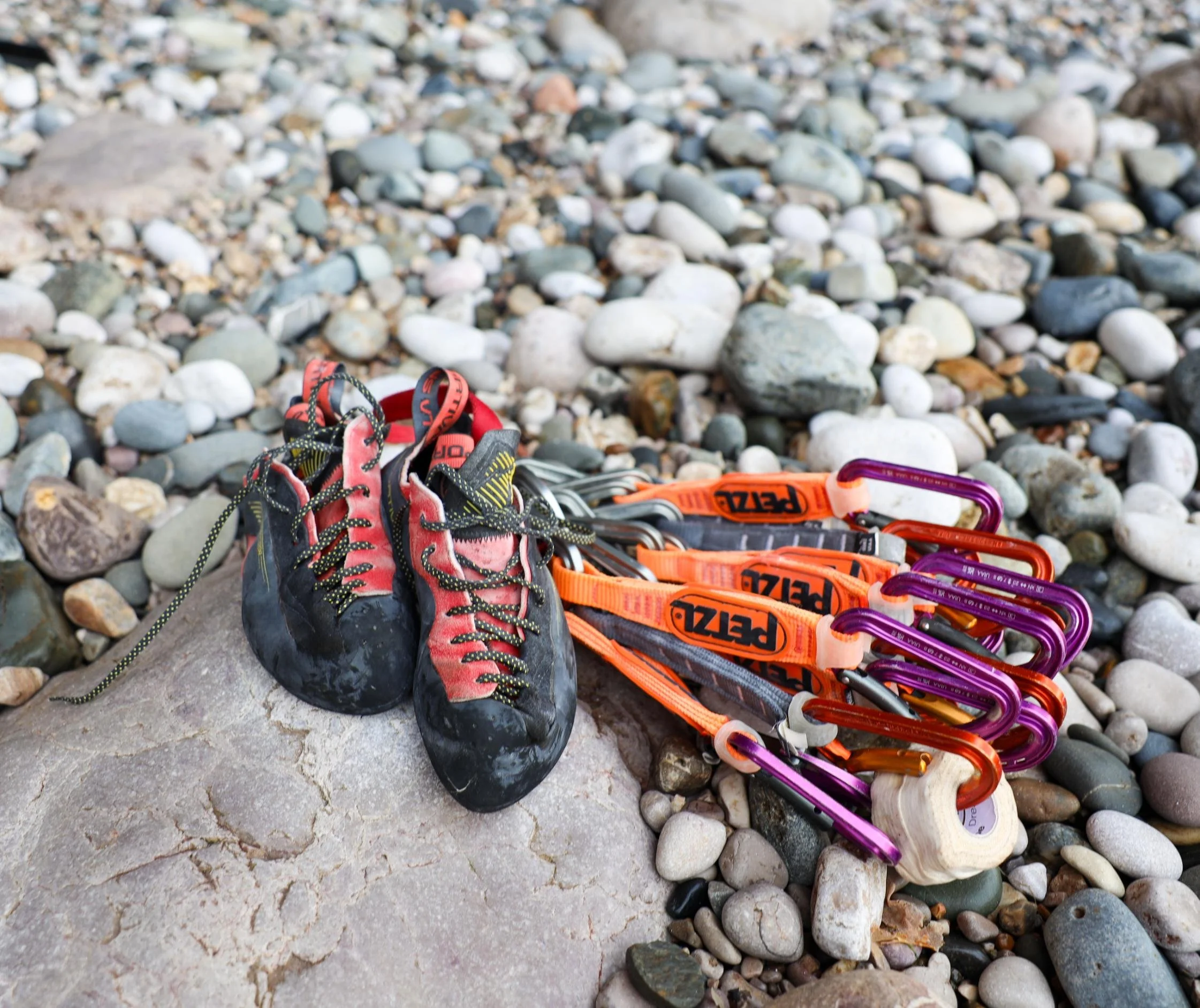 A pair of well-used climbing shoes, various colorful climbing gear including carabiners and straps, all placed on a rocky pebble beach.