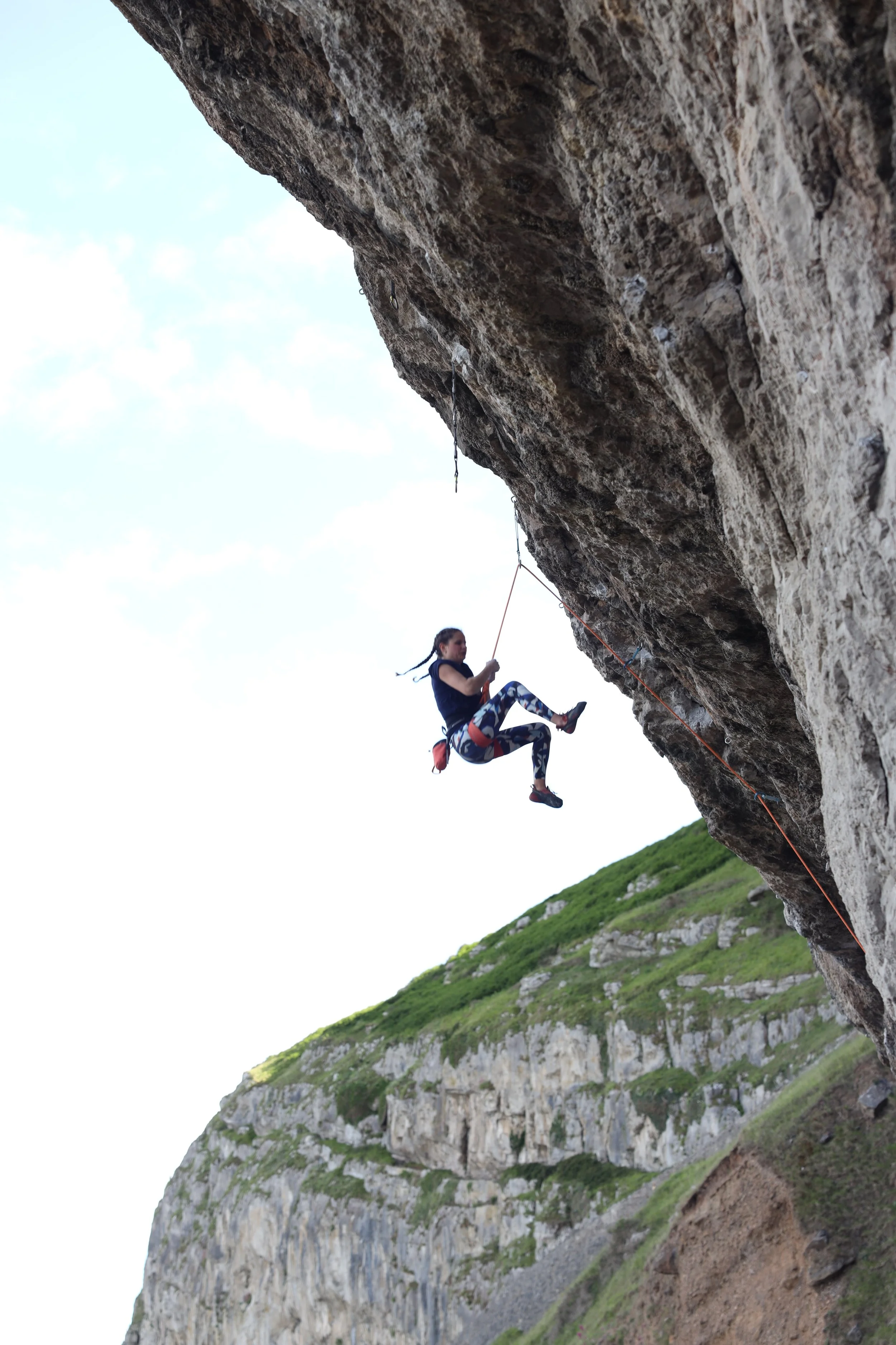 A woman bouldering on a steep outdoor rock face with green hills in the background.