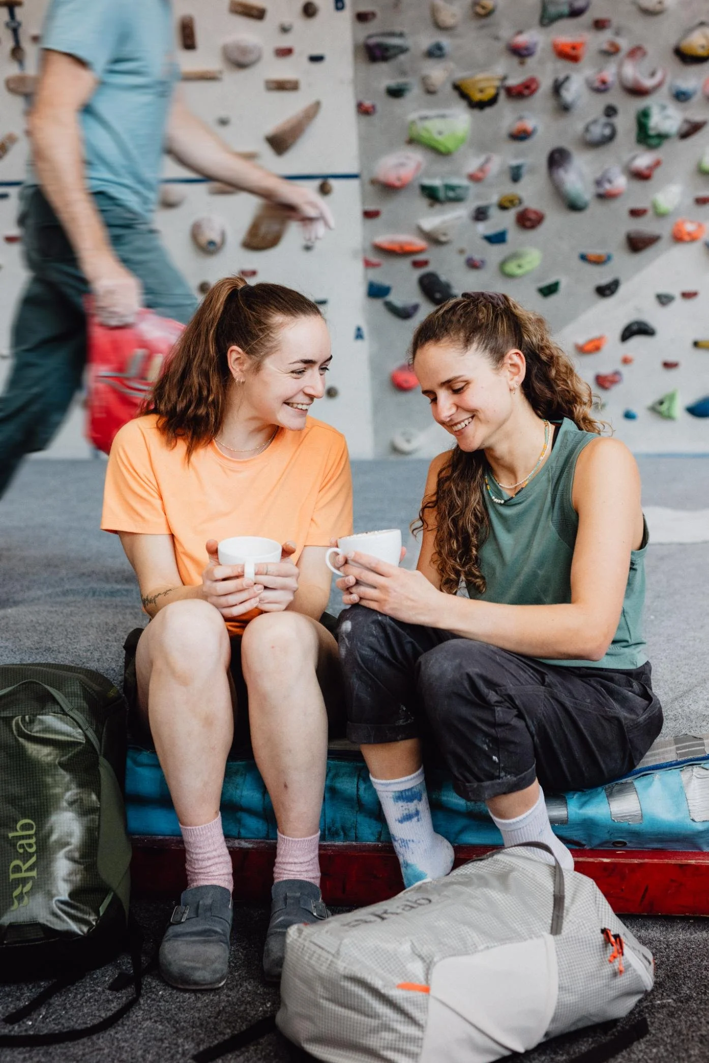 Two young women sit on a bench in an indoor climbing gym, smiling and holding mugs. Behind them is a climbing wall with colorful holds, and nearby are backpacks and gear on the floor.