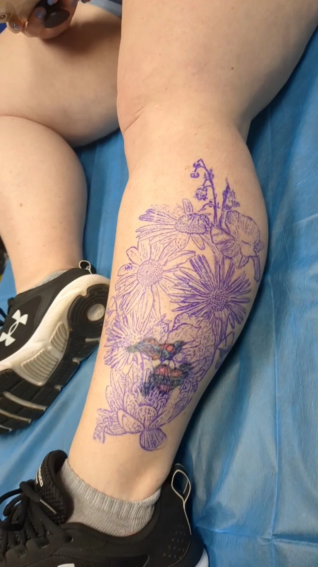 A person's leg with a tattoo outline of flowers, bees, and a woman's face, lying on a blue medical sheet near an Under Armour sneaker.
