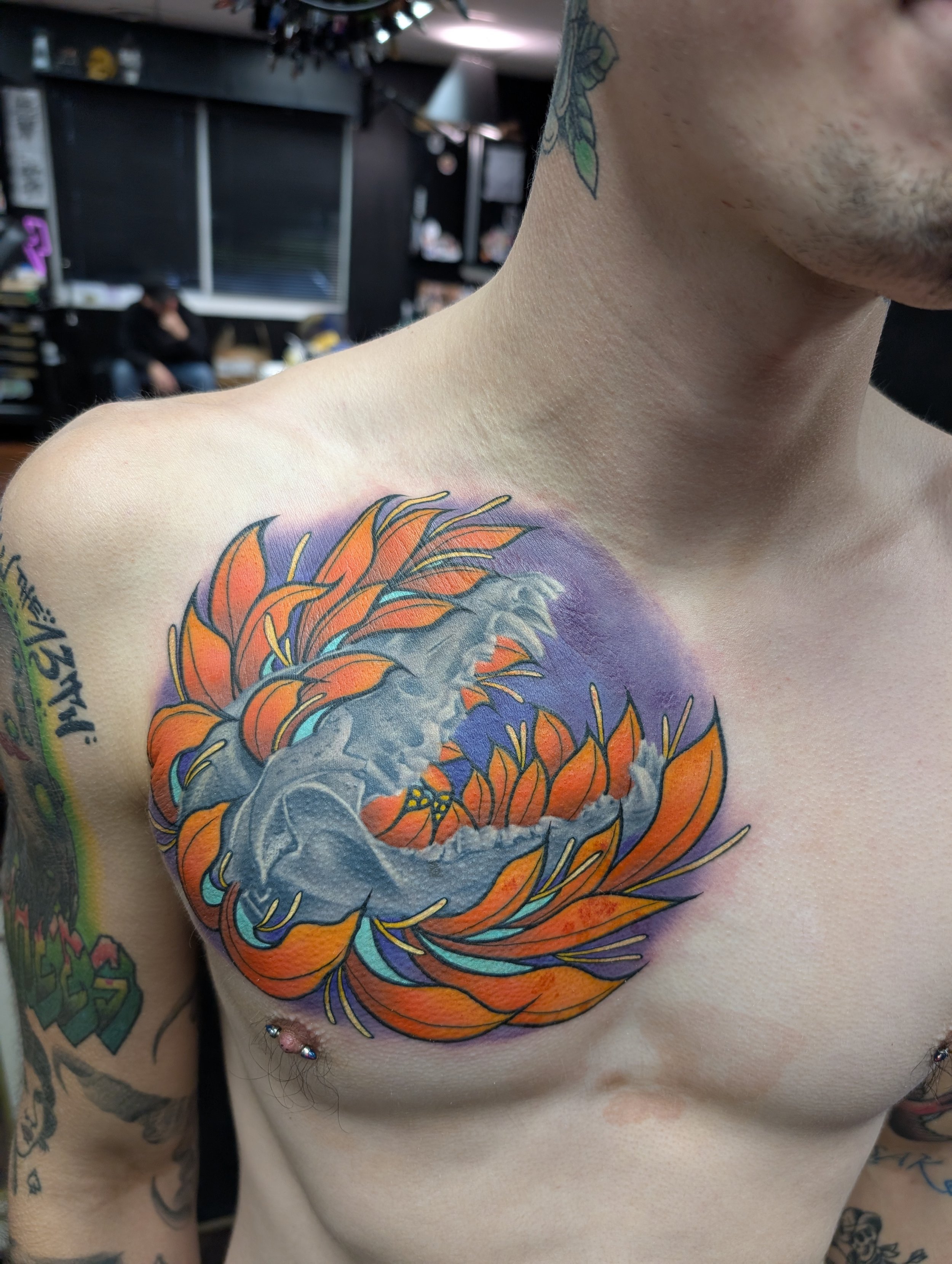 Close-up of a person's shoulder with a colorful tiger tattoo, showing orange, black, gray, purple, and yellow details, in a room with black walls and tattoos on other parts of the body.