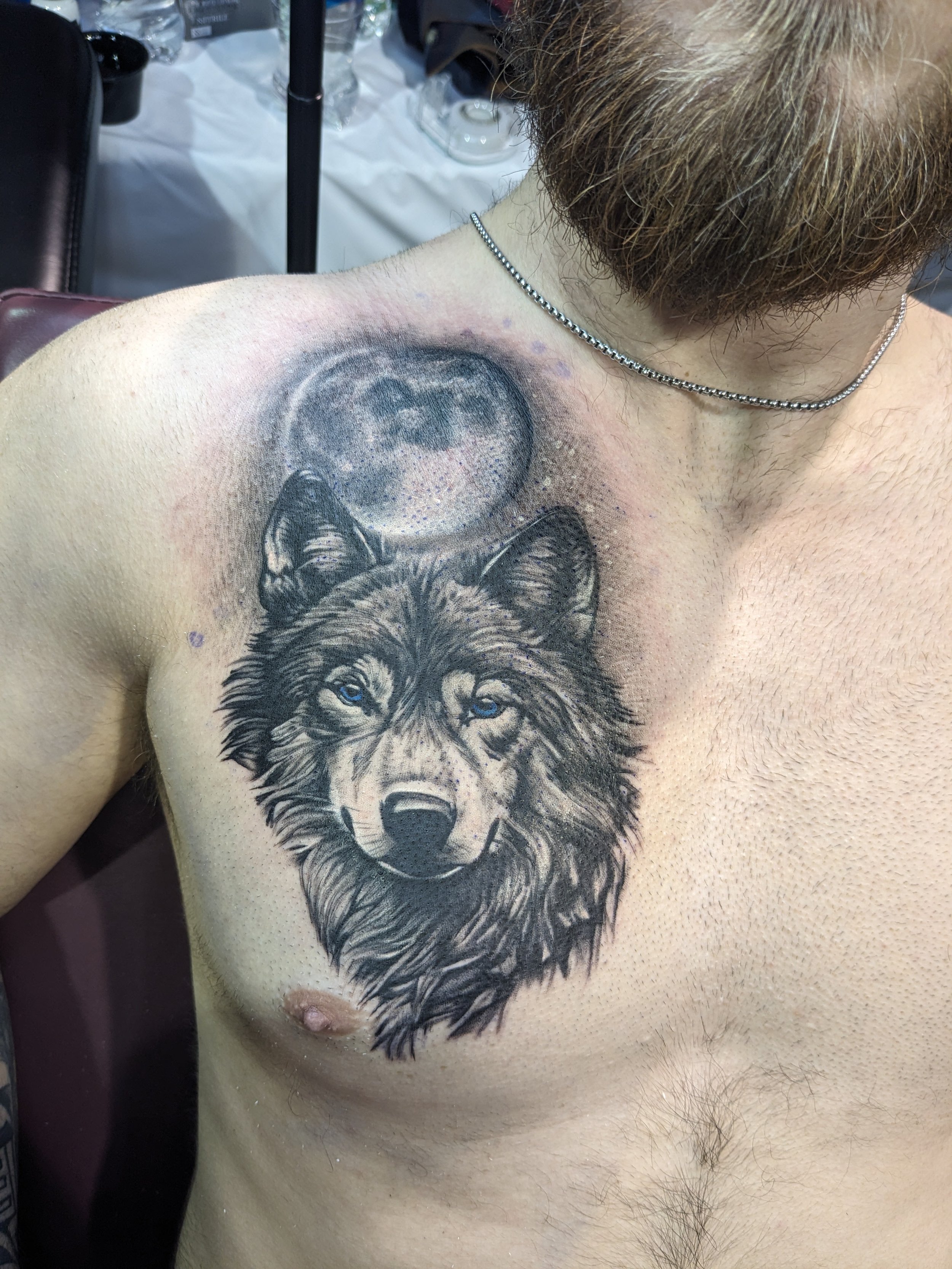A person with a partially completed tattoo of a wolf's face on their upper chest and shoulder. The tattoo features a detailed, realistic portrait of a wolf with piercing blue eyes, and there is a moon in the background. The person has light skin, che