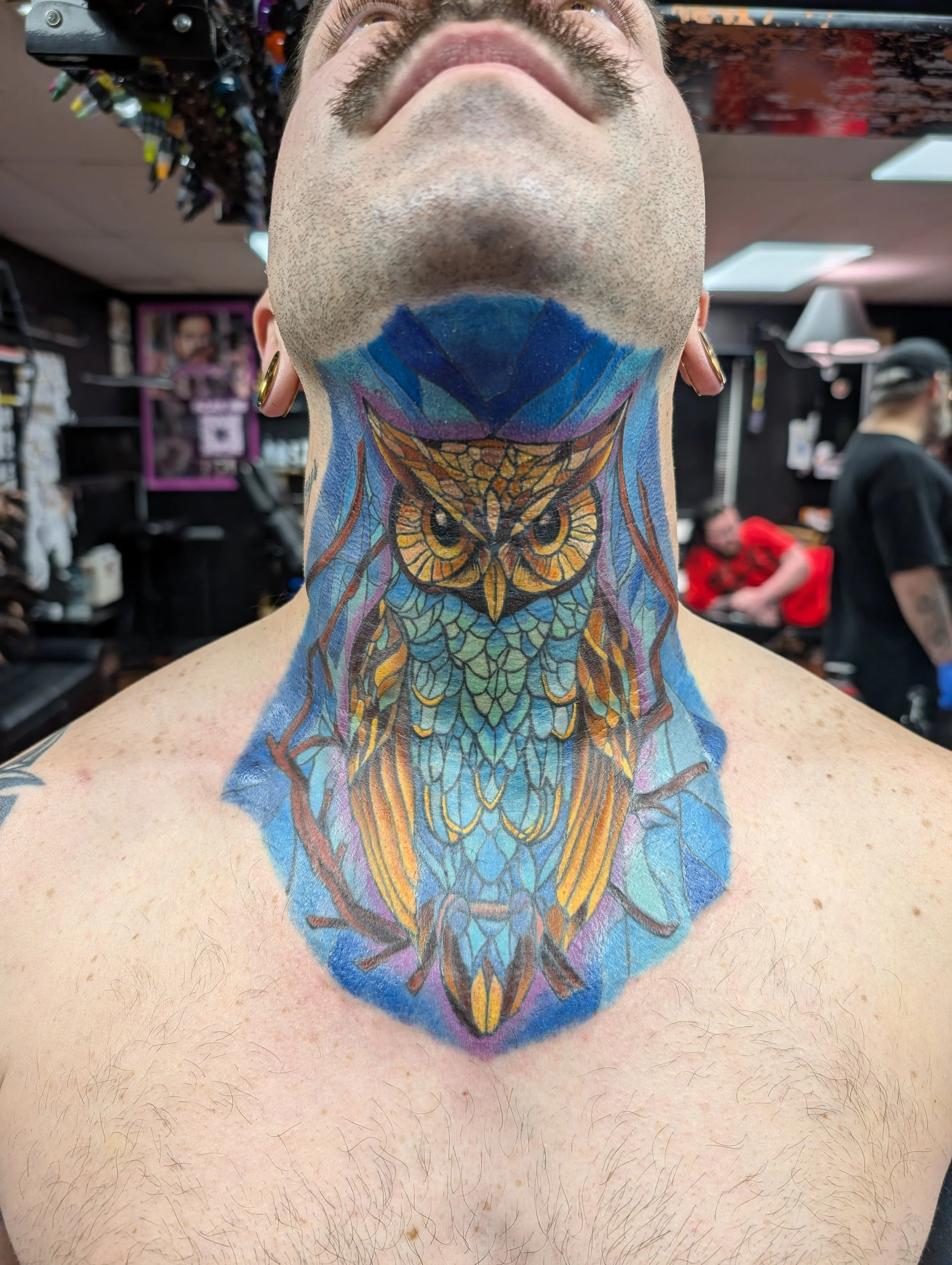A person with tattoos on their neck and chest, showing an owl design on their neck and chest, with a barber shop or tattoo studio background.