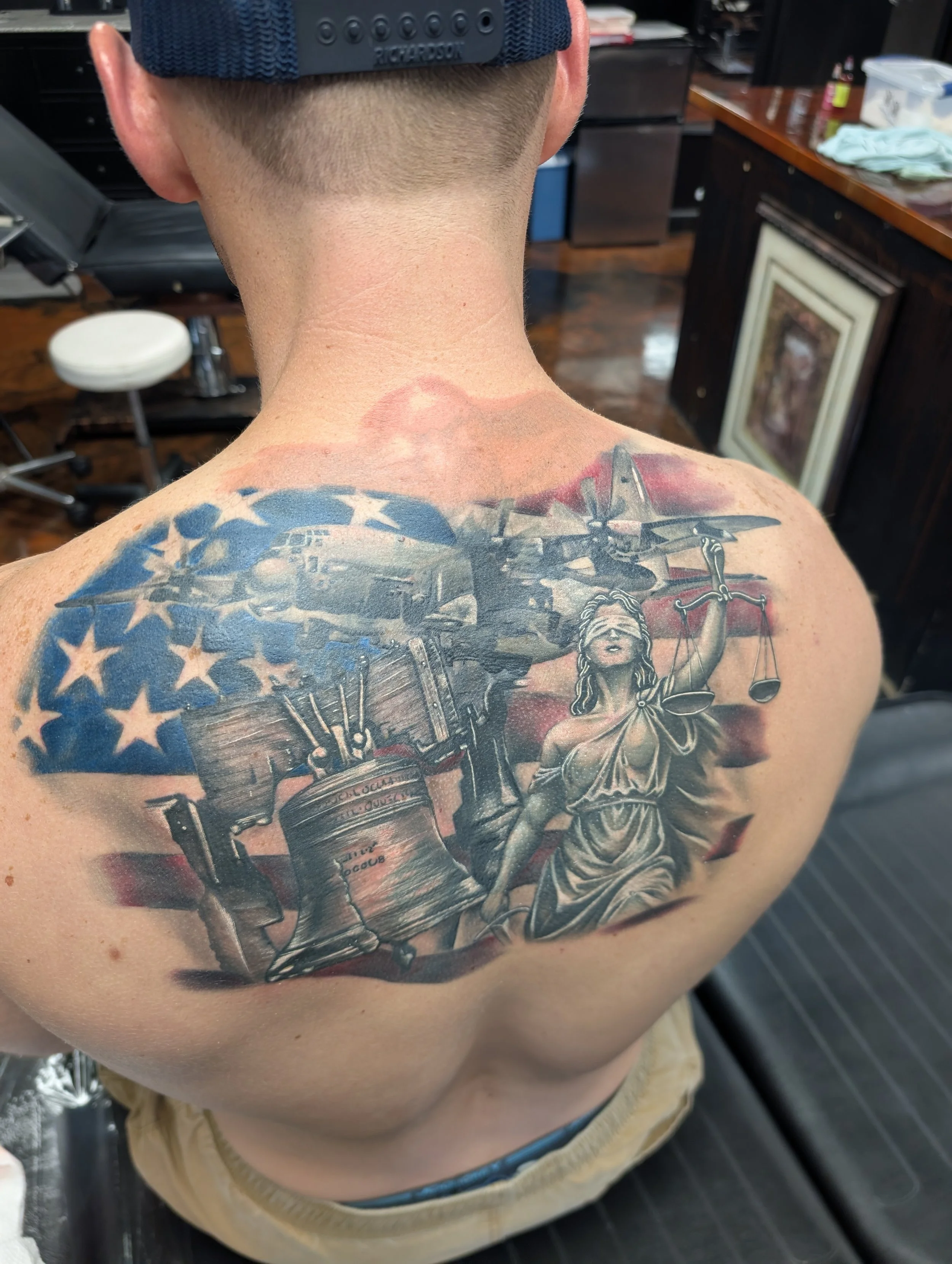 The back of a person's head and upper back, showing a tattoo of a woman holding scales, with a spray paint can, and a background of a tattoo studio.
