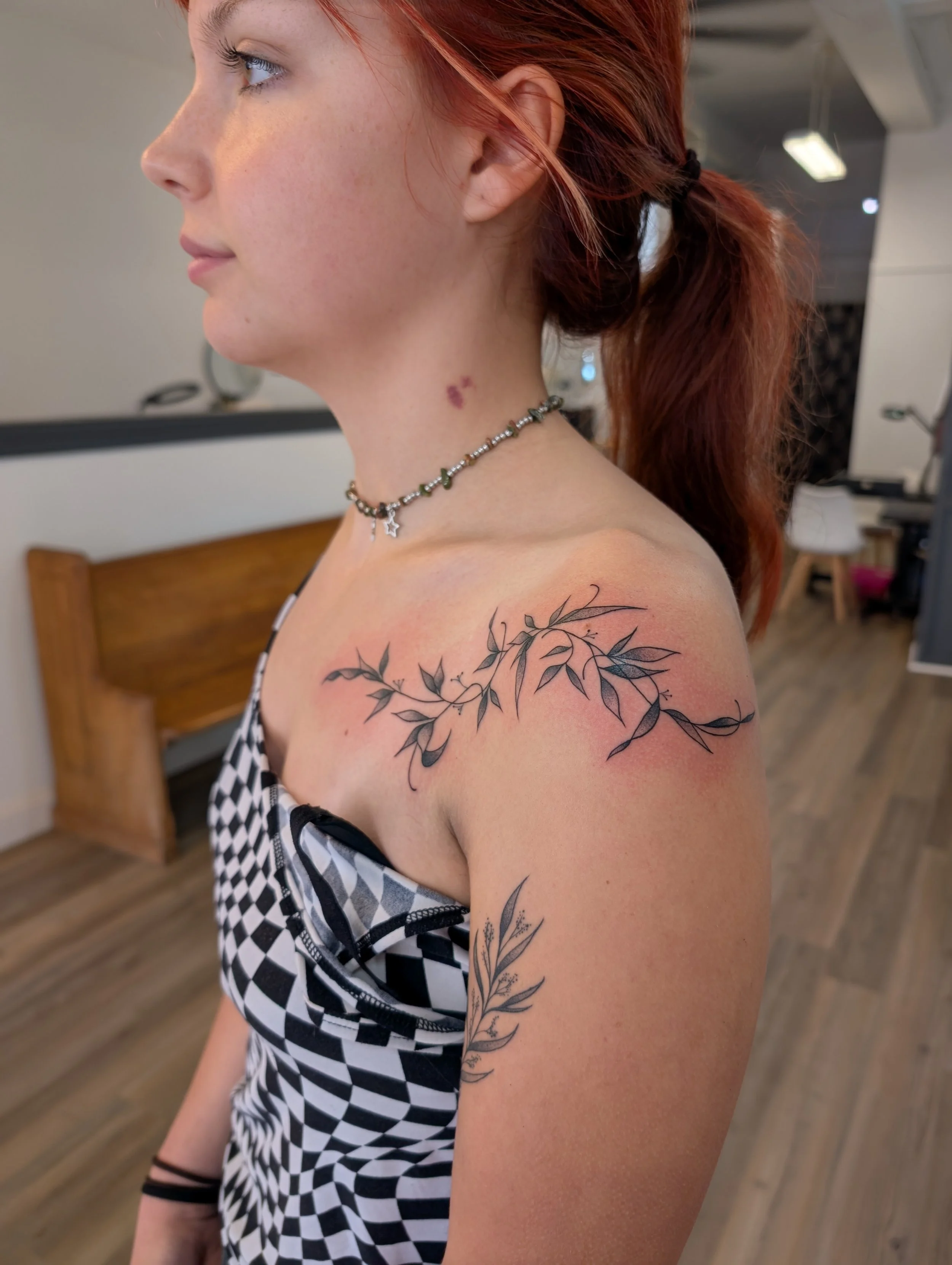 A woman with red hair ties in a ponytail, wearing a checkered black and white top and a star necklace, has a tattoo of eucalyptus branches on her shoulder and a purple birthmark on her neck, standing indoors.