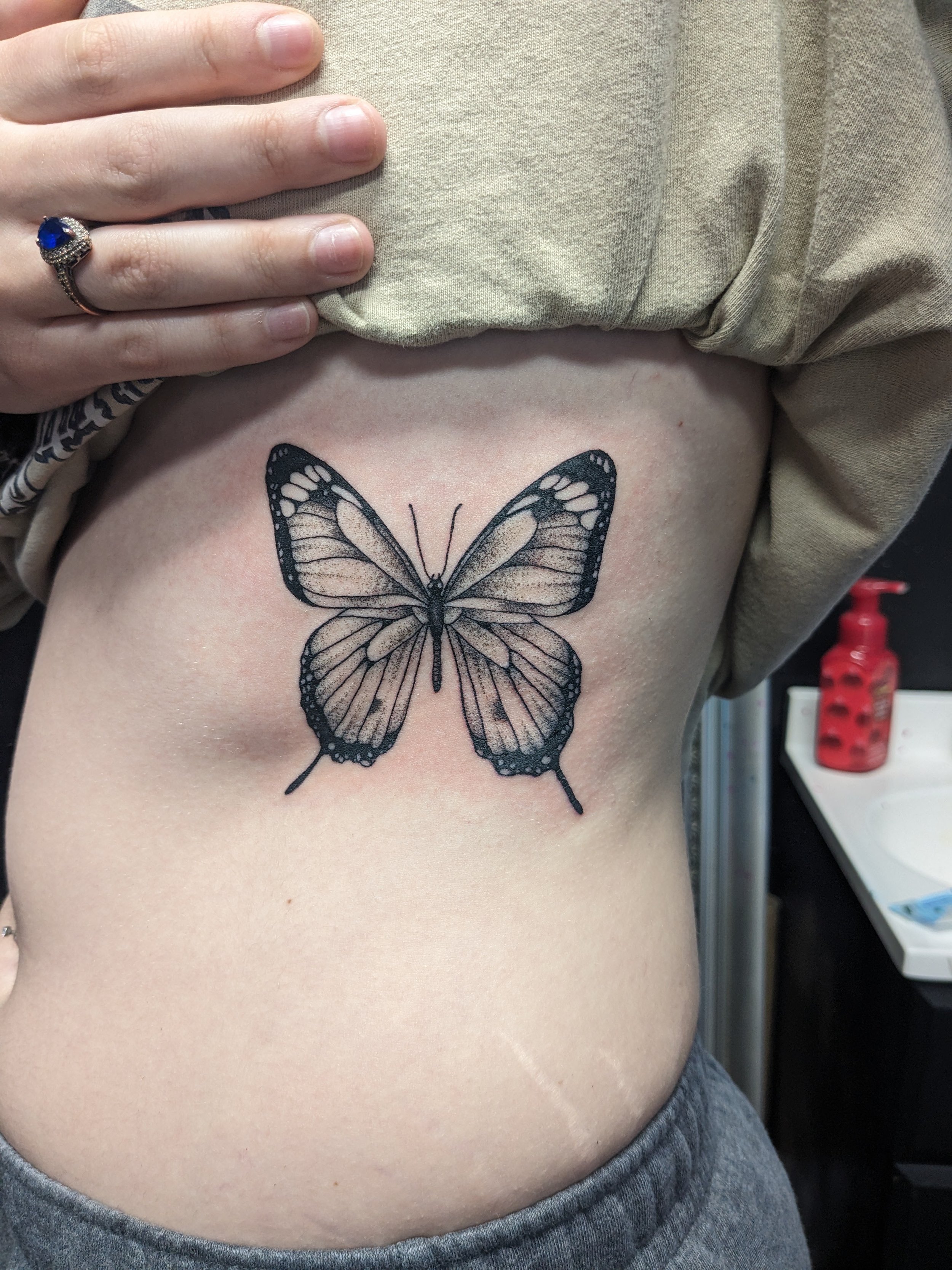 A person's side torso with a butterfly tattoo, hand with a ring, and beige shirt.