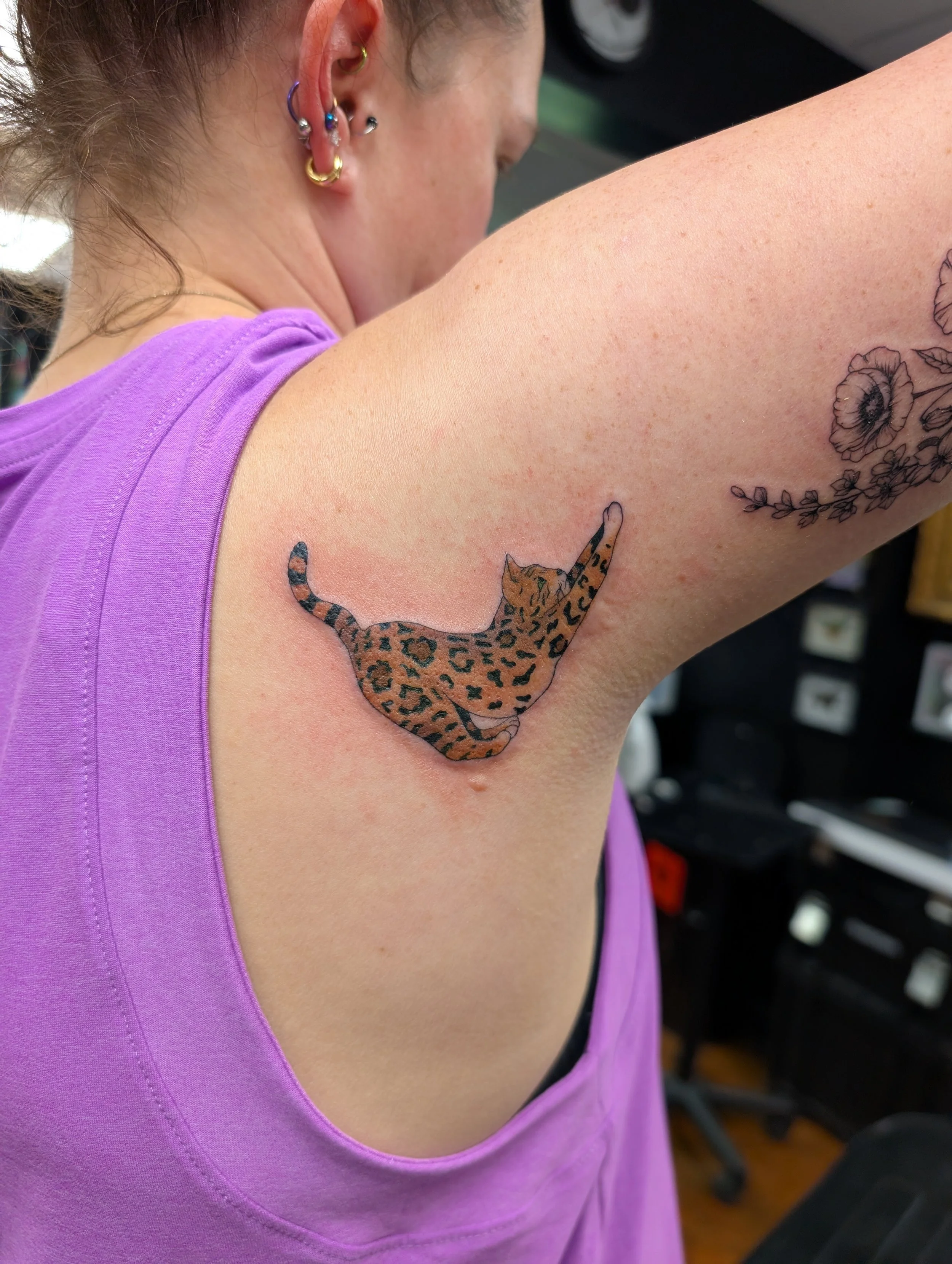 Close-up of a woman's arm with tattoos, including a leopard-print cat and flowers, wearing a purple top, showing several earrings in her ear.