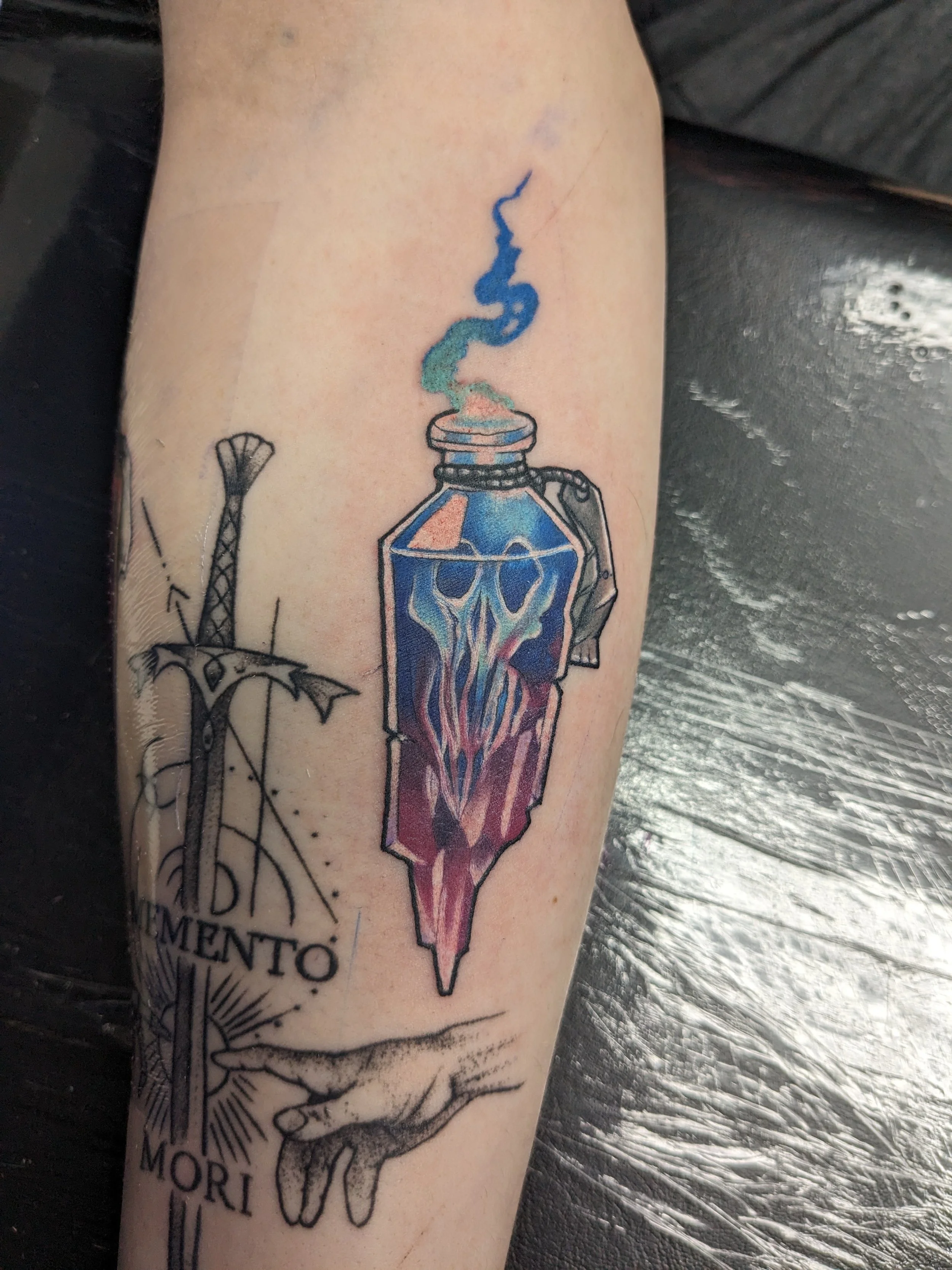A tattoo on a person's arm featuring a bloody dagger piercing a blue, stylized crystal. The tattoo also includes part of a text or design with black lines and shading.