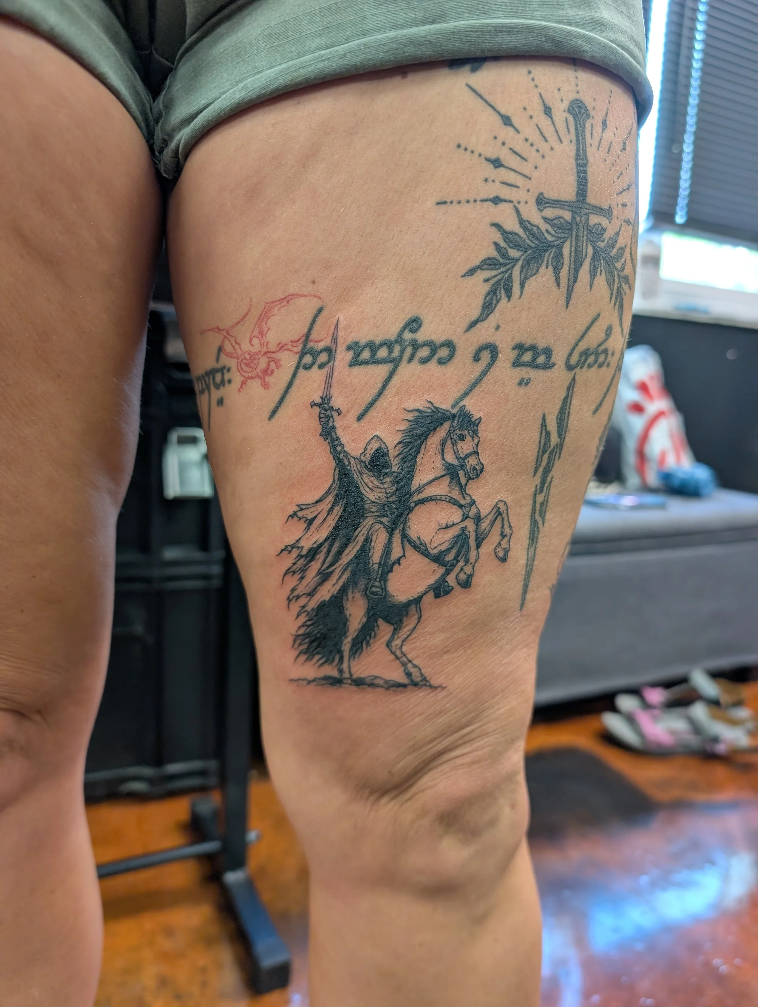 Tattoo of a sword crossed with a spear, surrounded by rays and leaves, with a rider on a horse holding a sword in the lower part of the thigh.