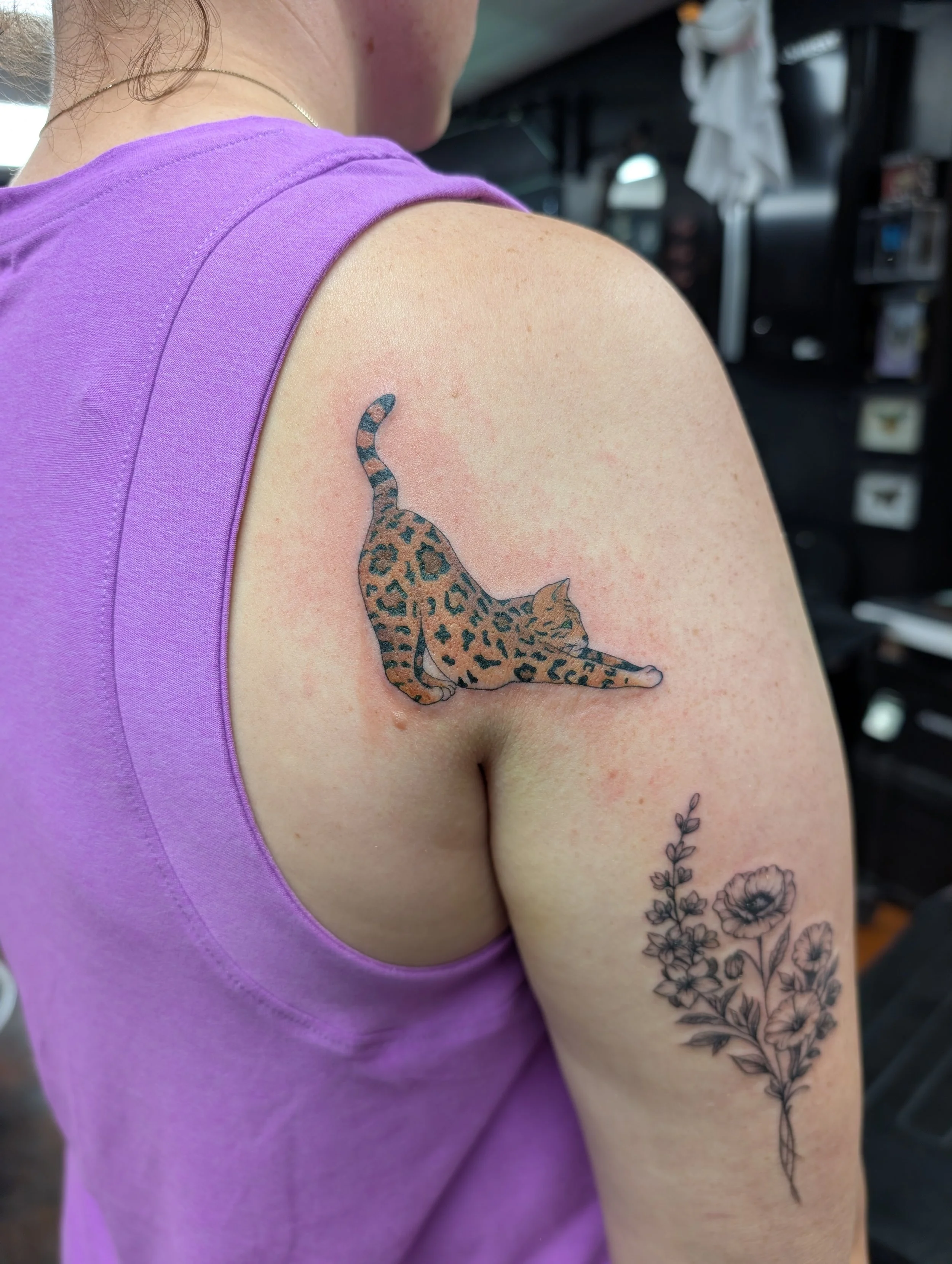 Close-up of a woman's shoulder and arm with a leopard-print cat tattoo on her upper arm and a floral tattoo on her forearm, wearing a purple sleeveless top.