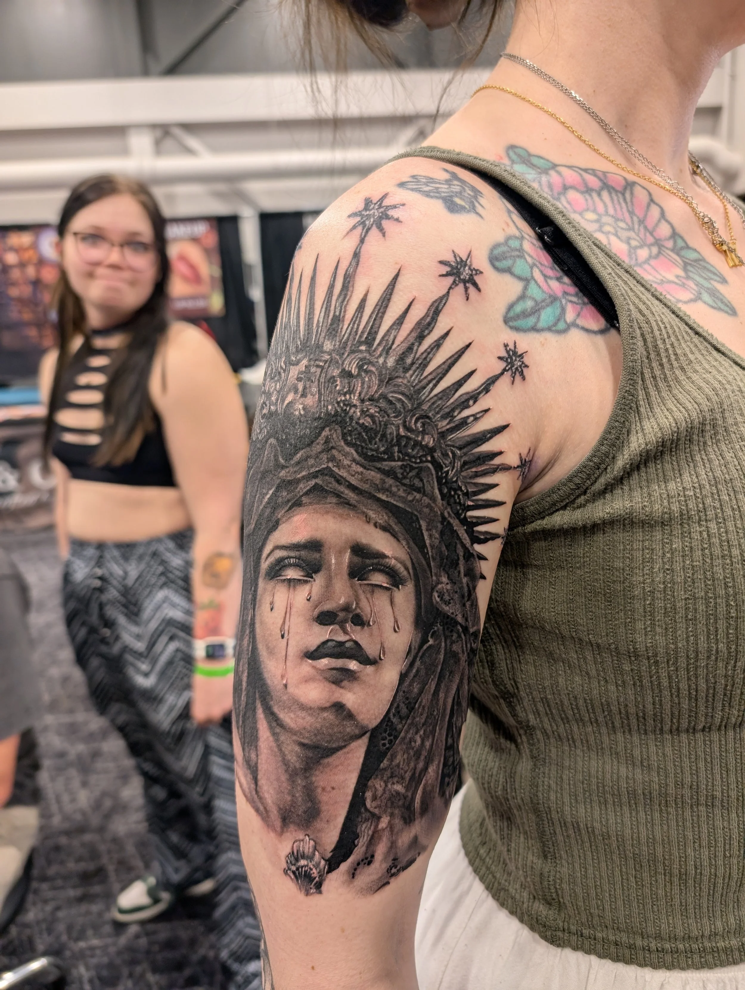 Close-up of a woman's arm with a large, detailed black and gray tattoo of a woman's face with tears, a crown, and various decorative elements. Another woman with glasses and a black sleeveless top is visible blurred in the background.