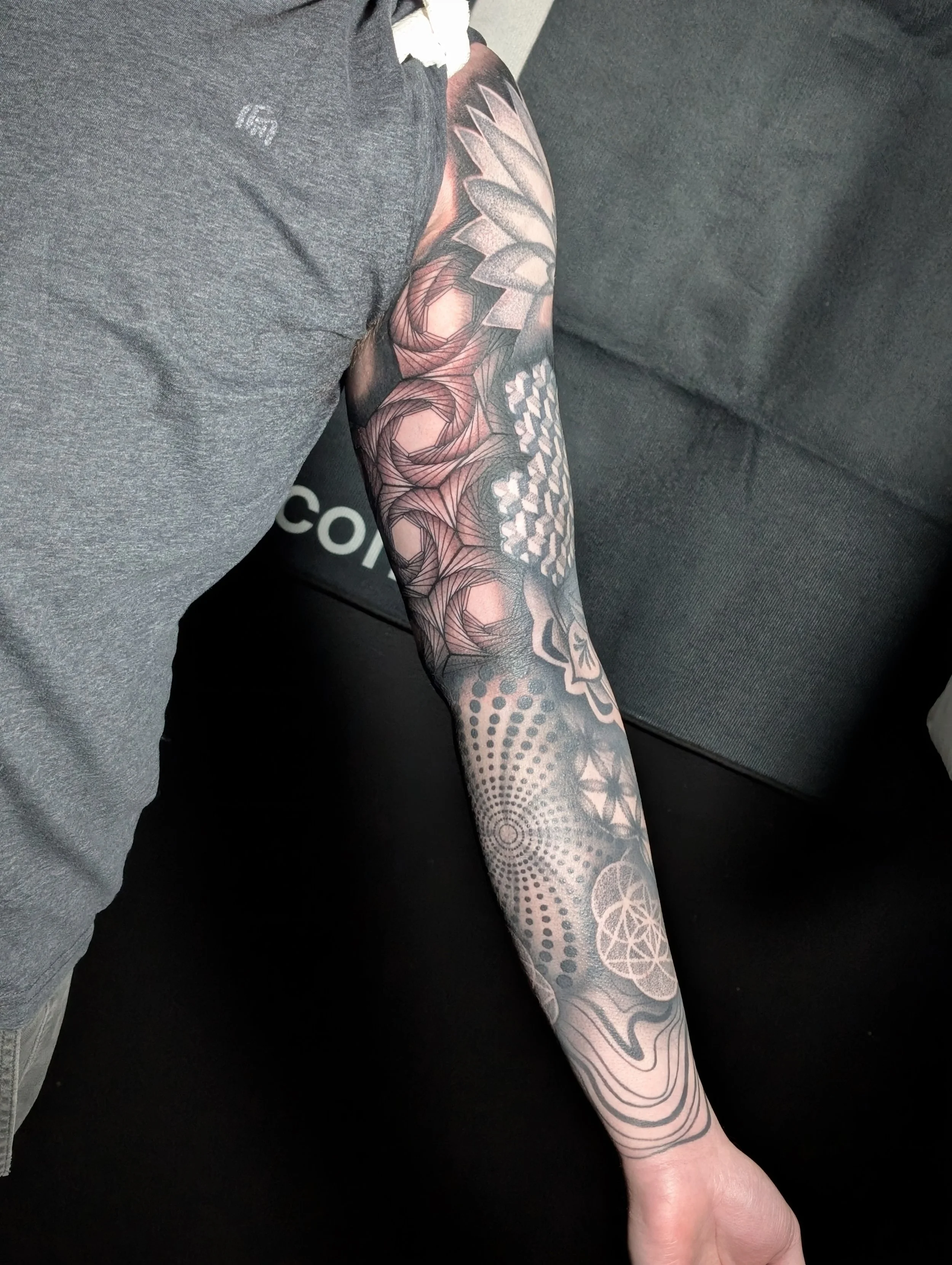 Tattooed arm with geometric patterns, roses, and other designs resting on a dark sofa.