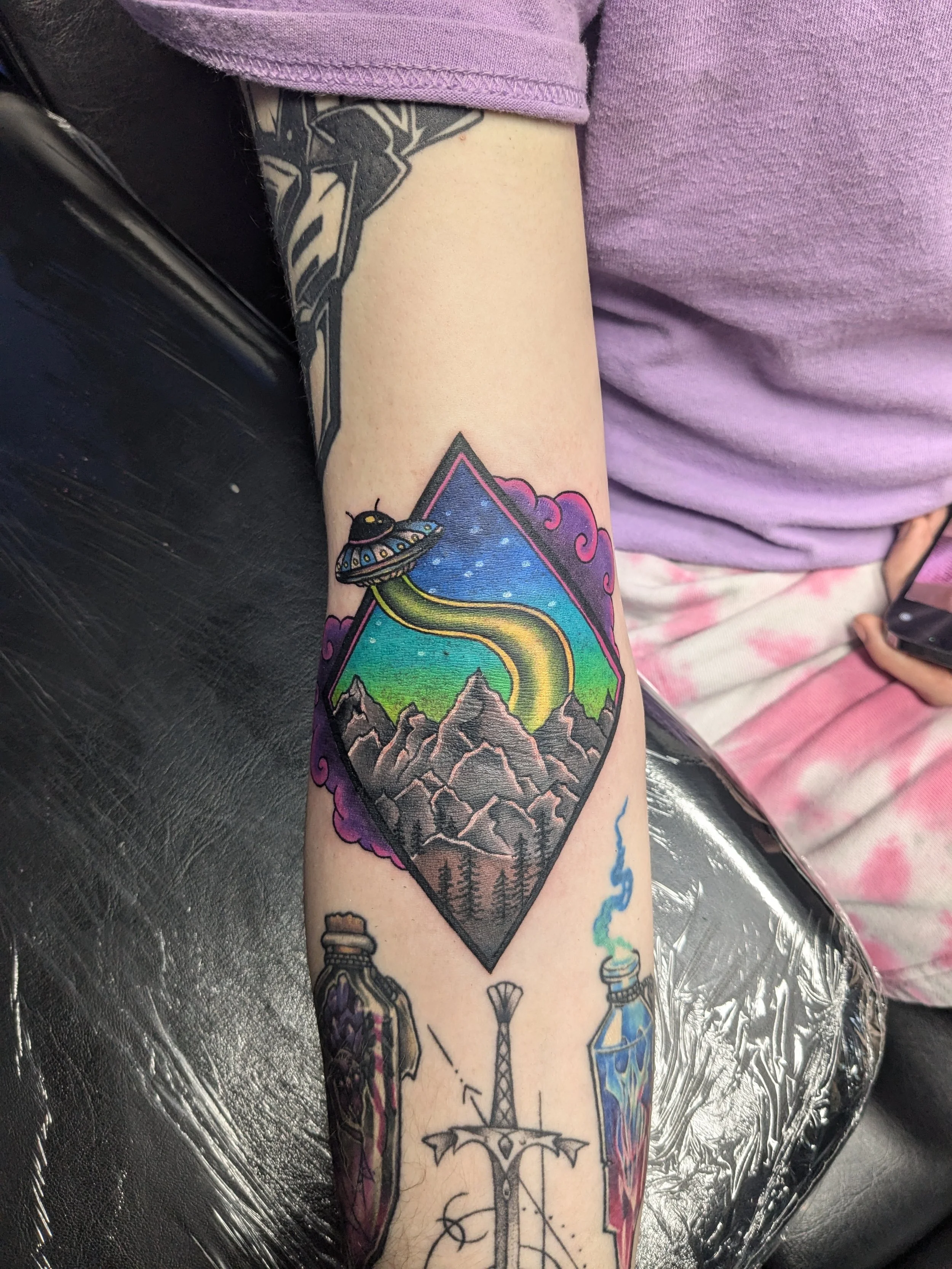 Colorful tattoo of a mountain landscape with a rainbow, clouds, and a UFO on the person's arm, with additional tattoo designs visible below.