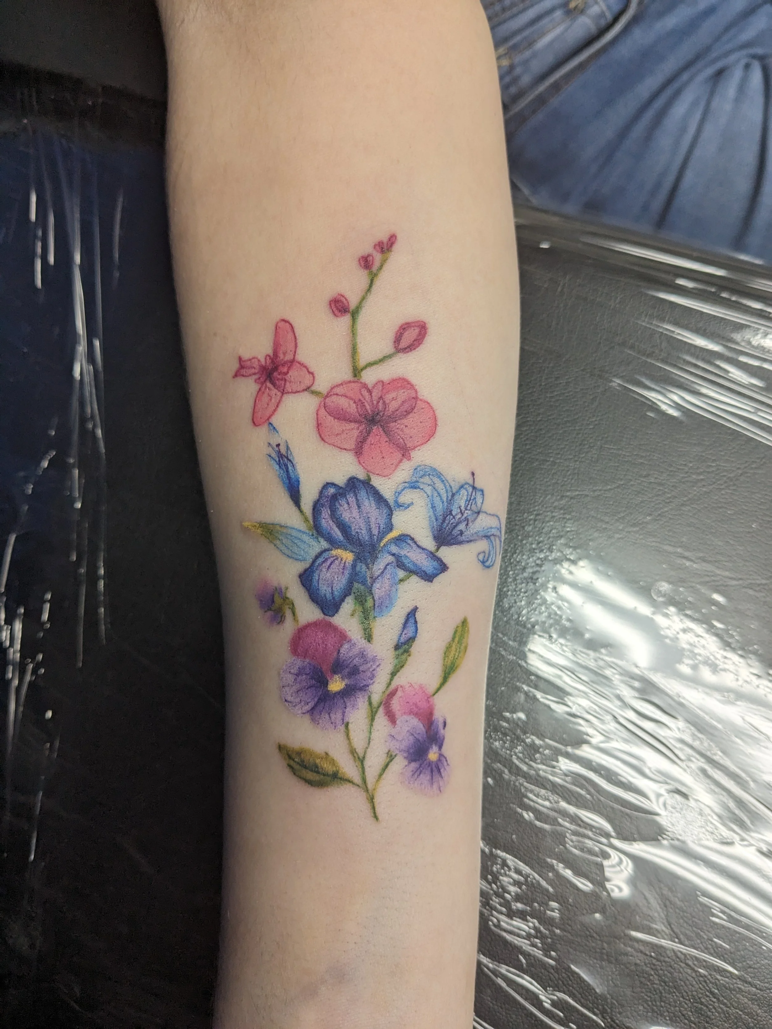 Close-up of a person's forearm with a colorful floral tattoo featuring pink, purple, and blue flowers and green stems.