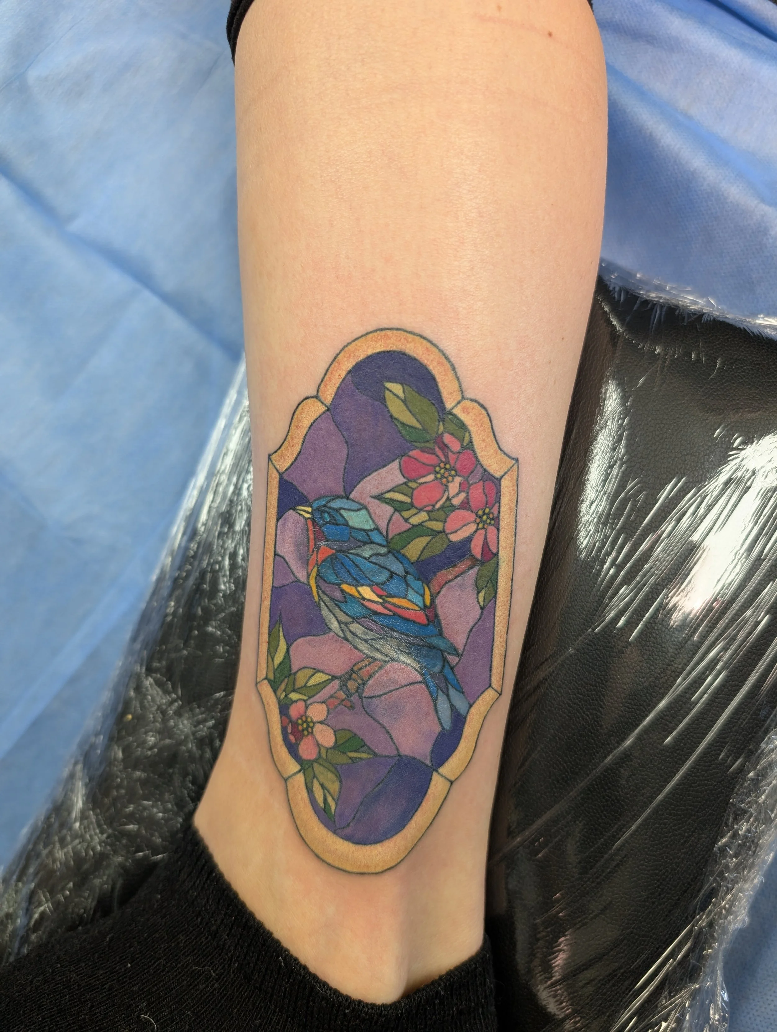 Colorful tattoo of a stained glass window with a bluebird and red flowers on a person's forearm.