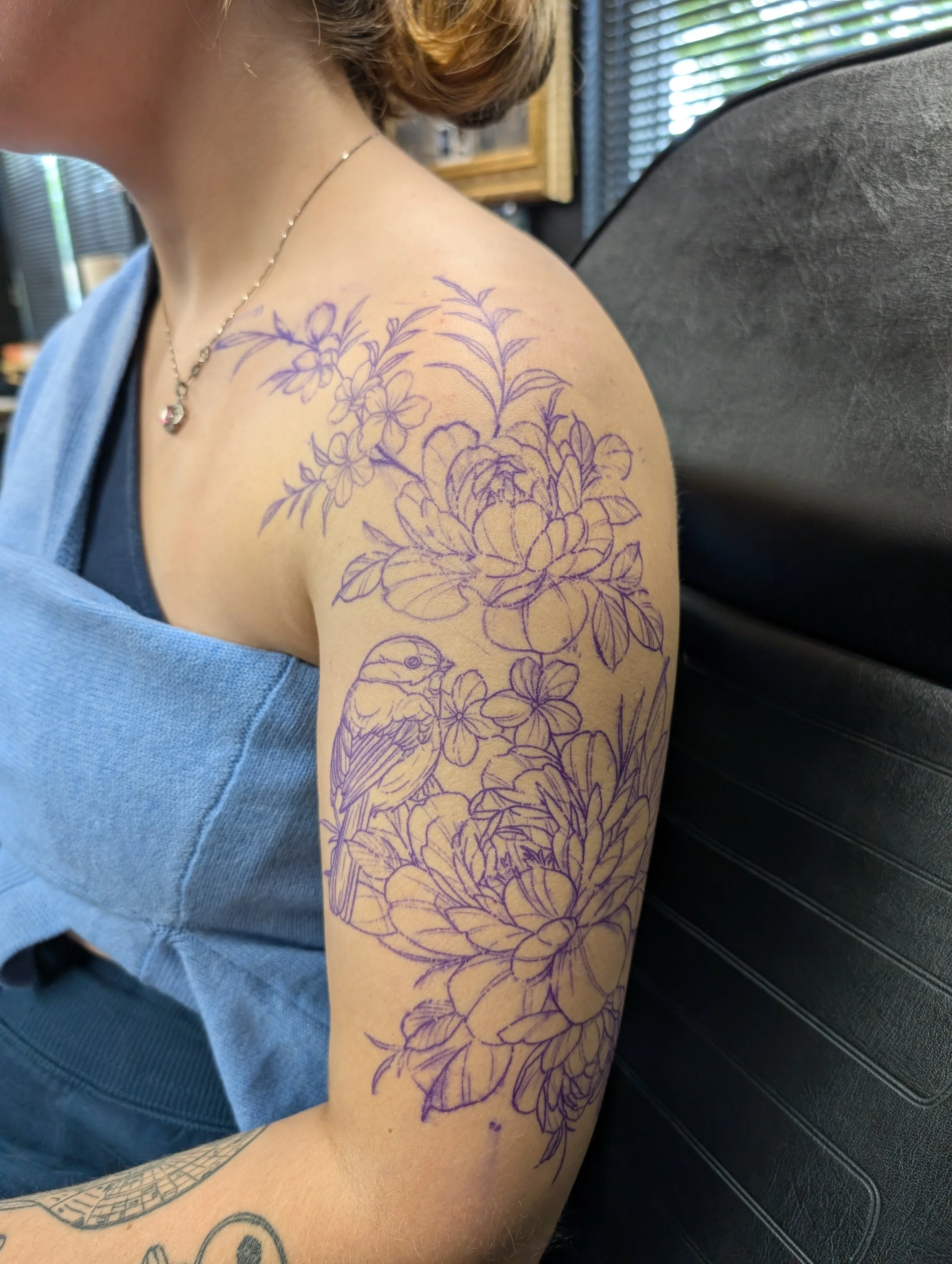 A person with a floral line drawing tattoo on their upper arm and shoulder, wearing a light blue top, sitting against a black surface with window blinds in the background.