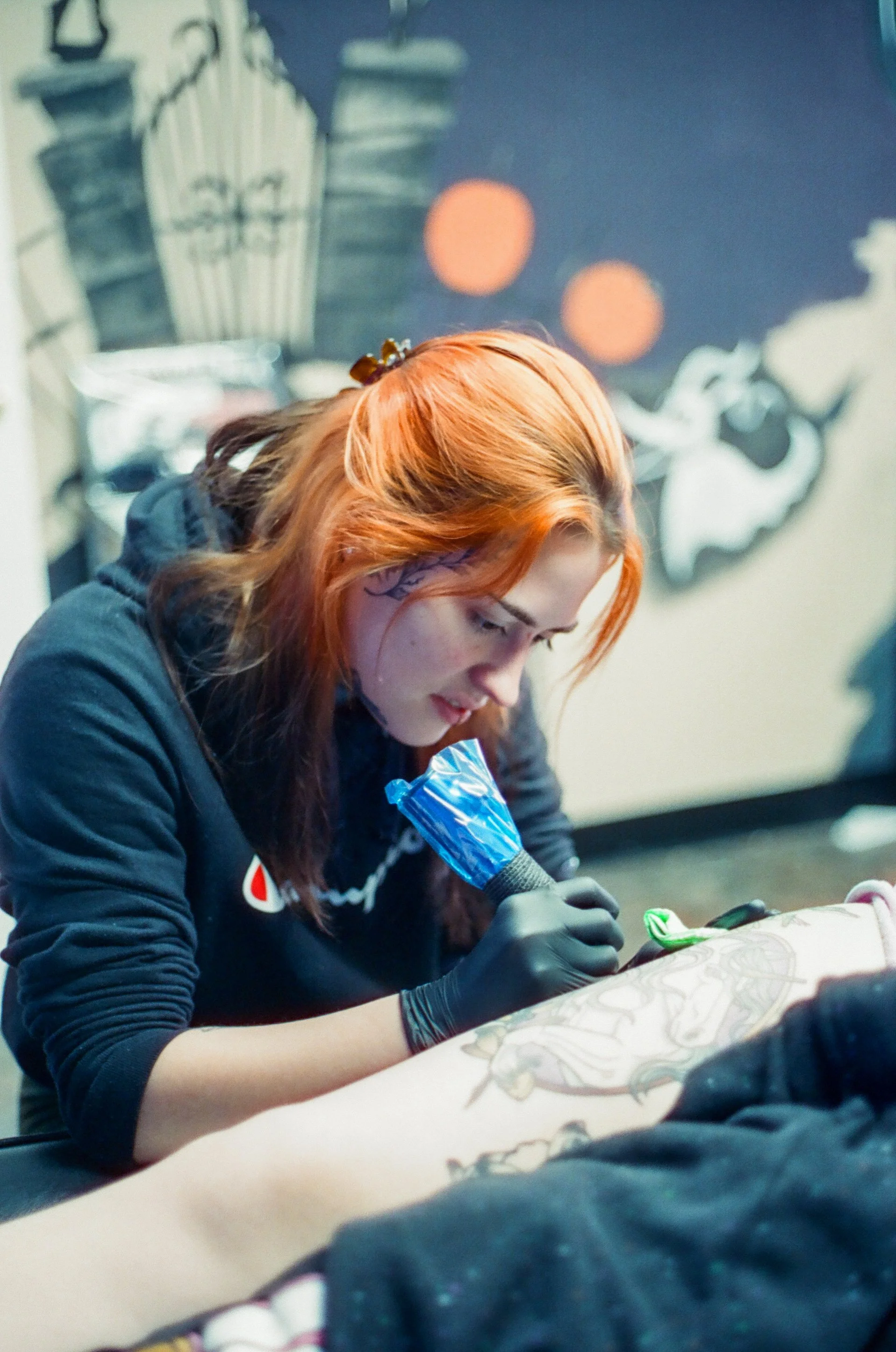 A woman with reddish hair, tattoos on her neck, wearing a black hoodie and gloves, tattooing on a person's back in a tattoo parlor.
