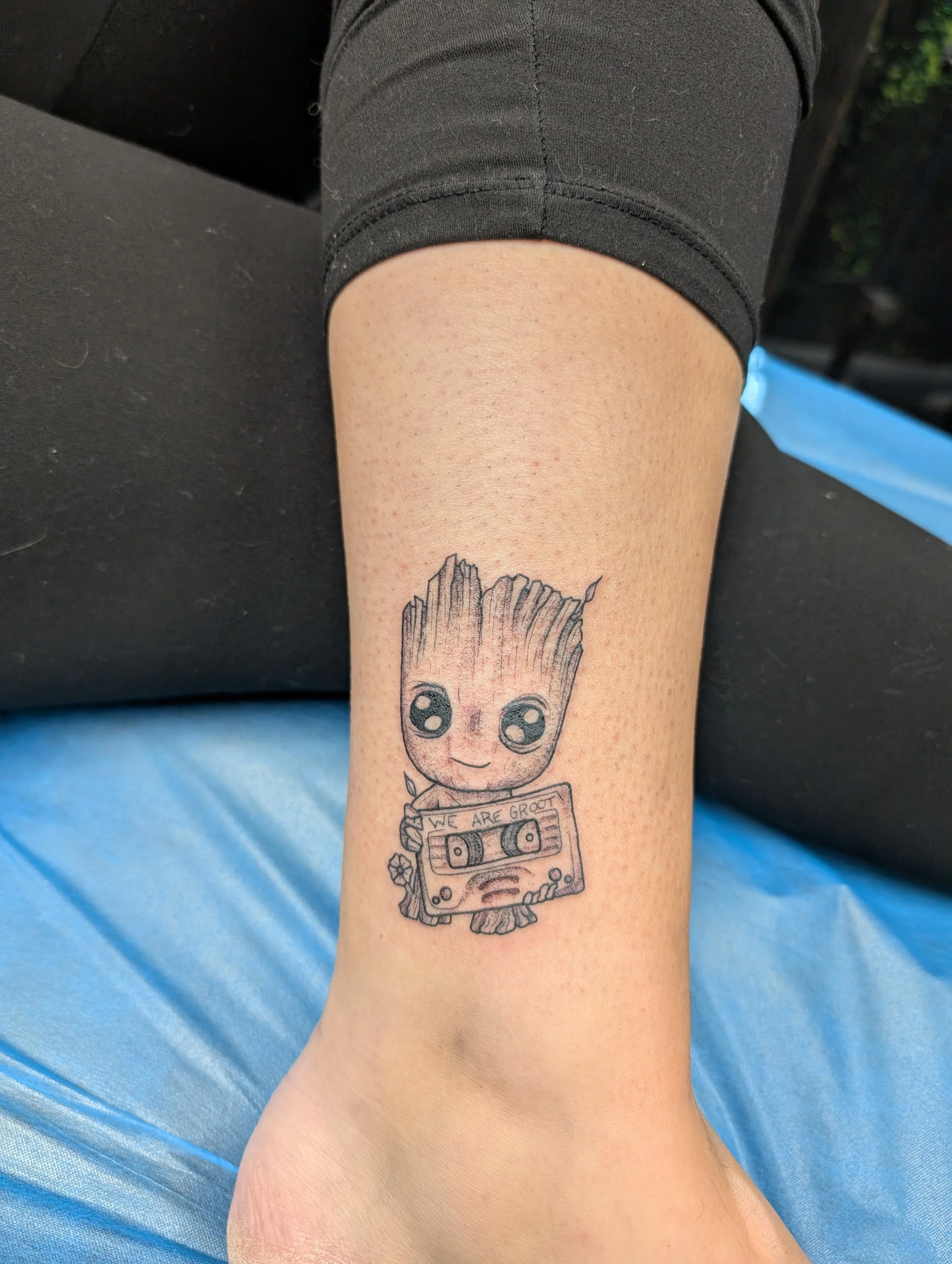 Close-up of a person's forearm with a tattoo of Baby Groot holding a cassette tape that says "We are Groot" with a small flower at the bottom.