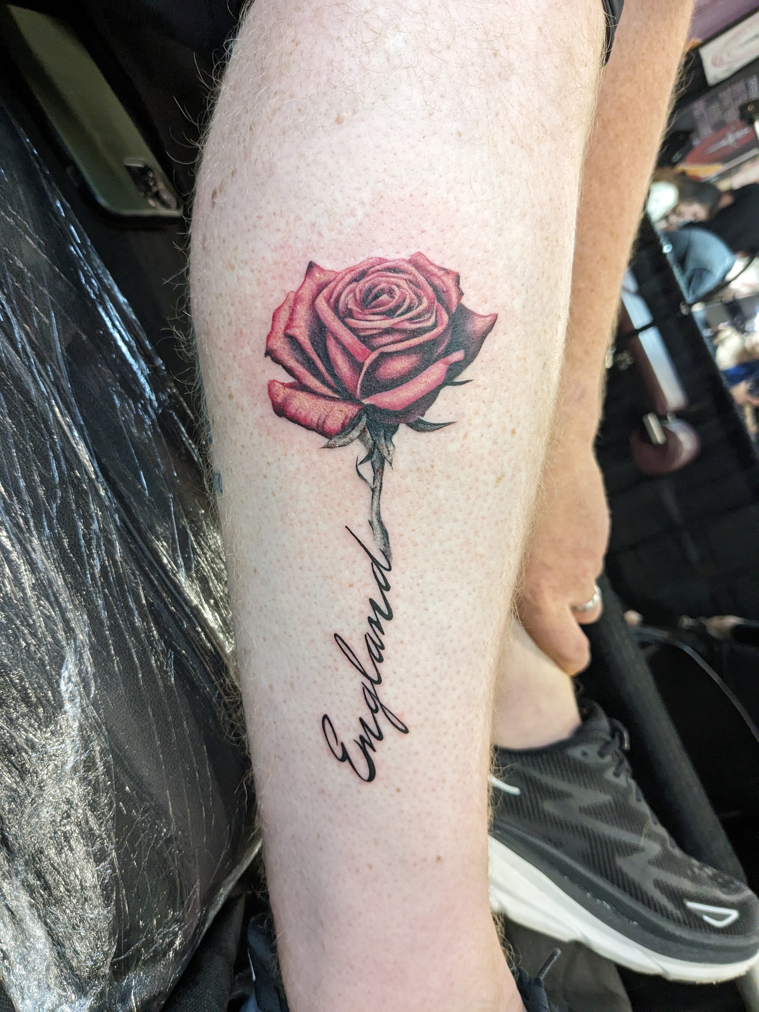 A close-up of a person's forearm with a tattoo of a pink and black rose and a cursive word underneath.