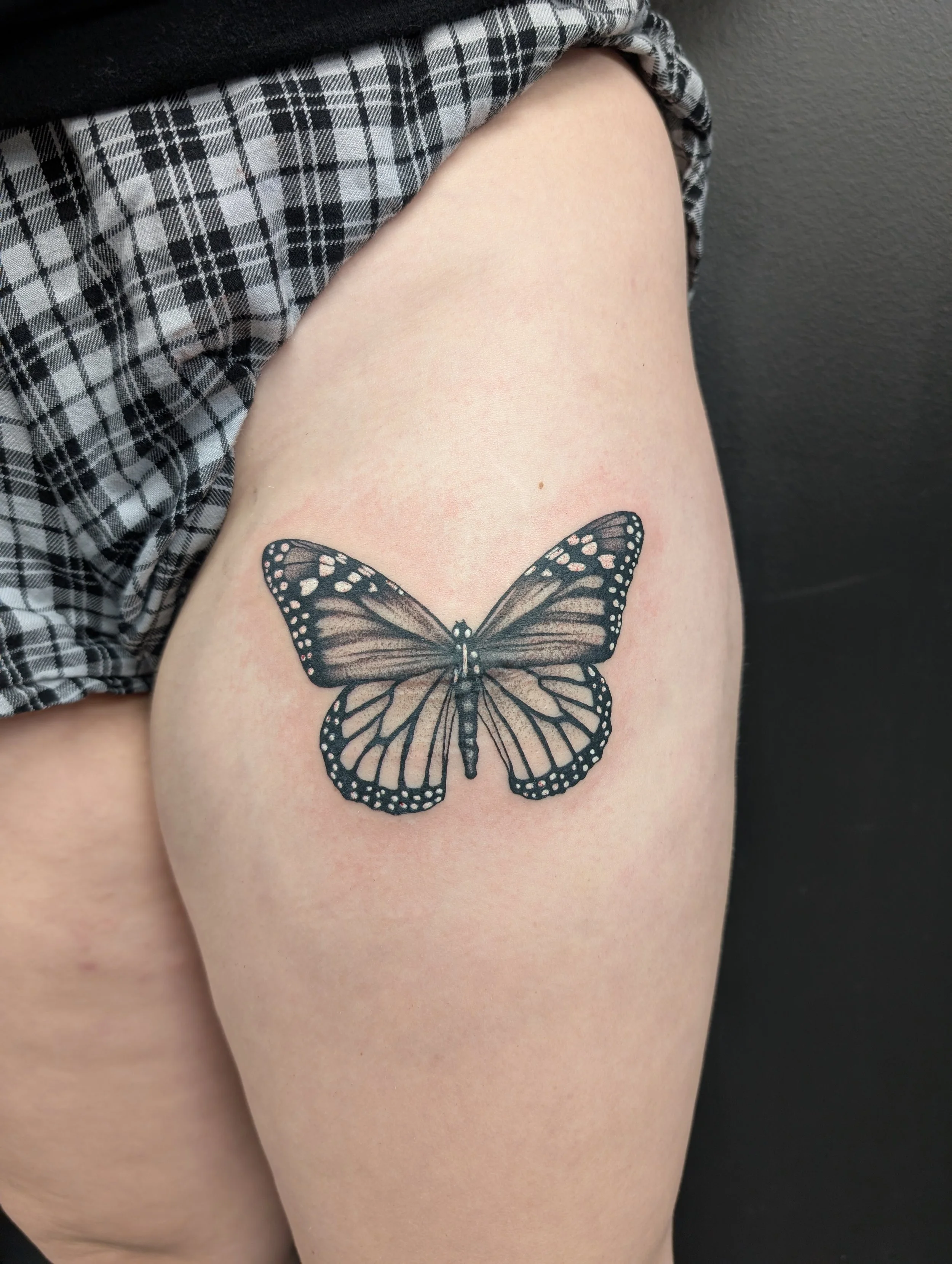 Close-up of a person's thigh with a butterfly tattoo below the knee, wearing a plaid garment and black top, against a gray background.