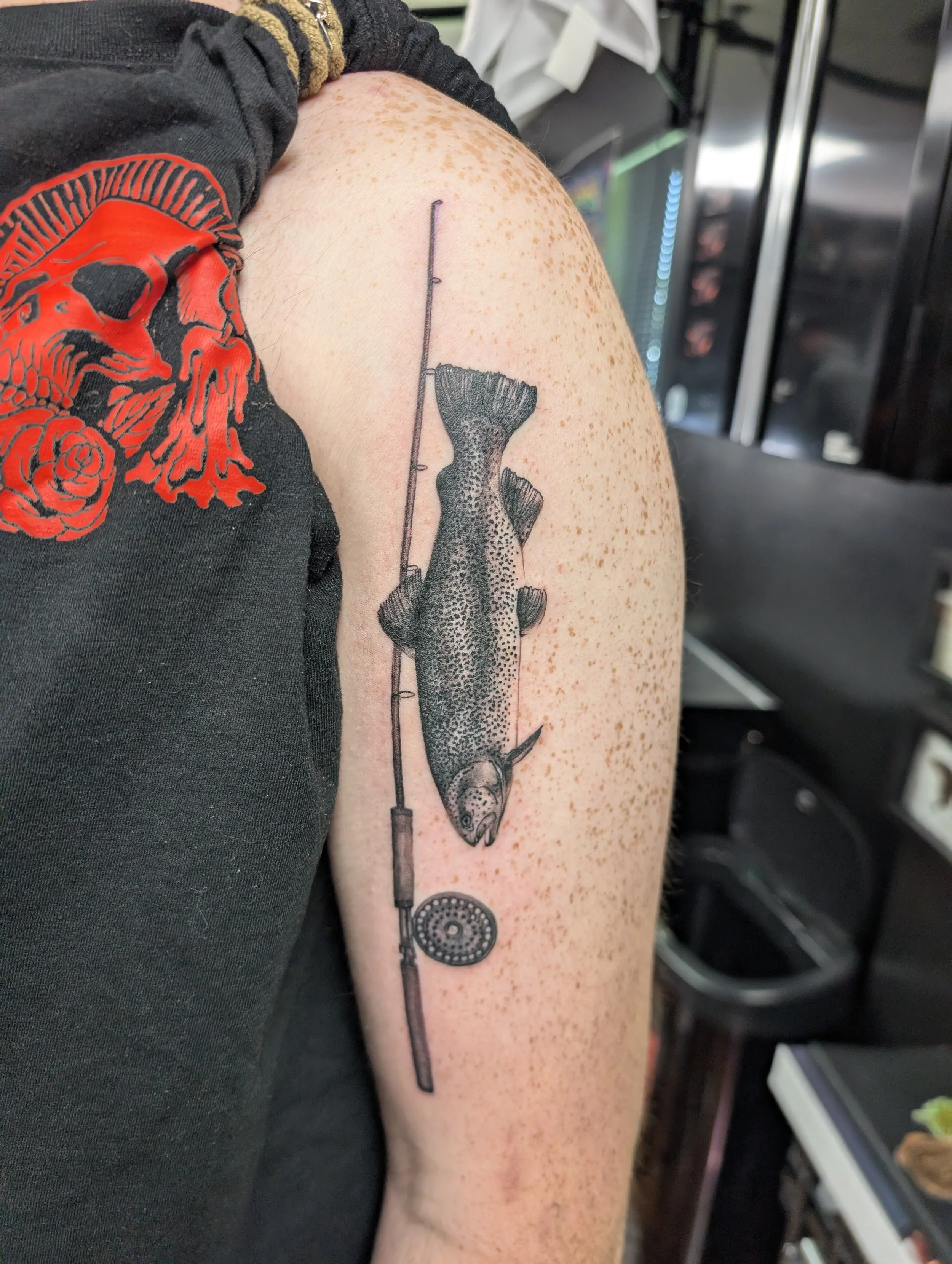 A tattoo on a person's upper arm depicts a fish hanging from a fishing line with a reel at the bottom, rendered in black ink with detailed shading.