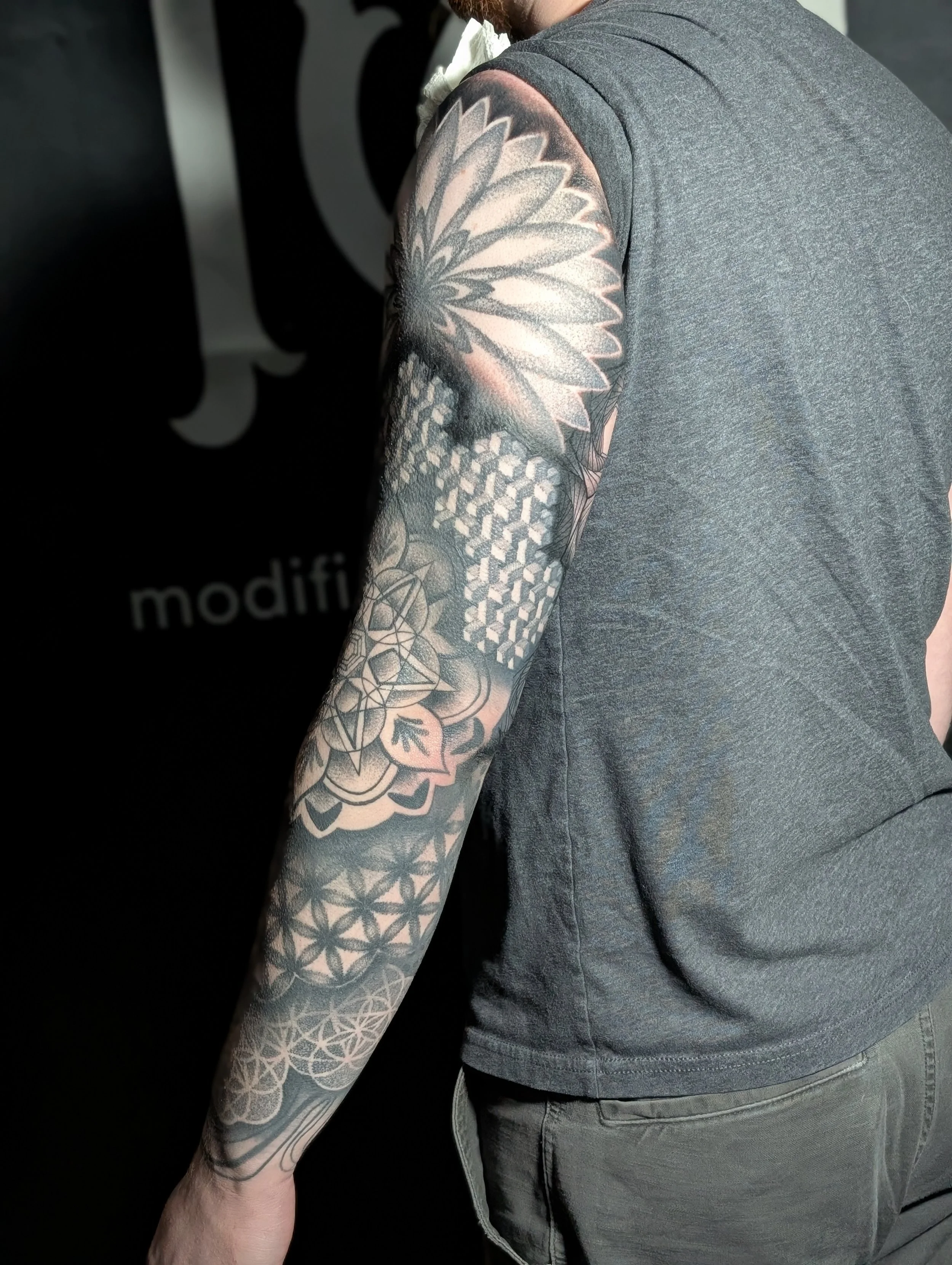 A man with tattoos on his arm in front of a black background with the word 'modifi' visible, wearing a gray t-shirt.