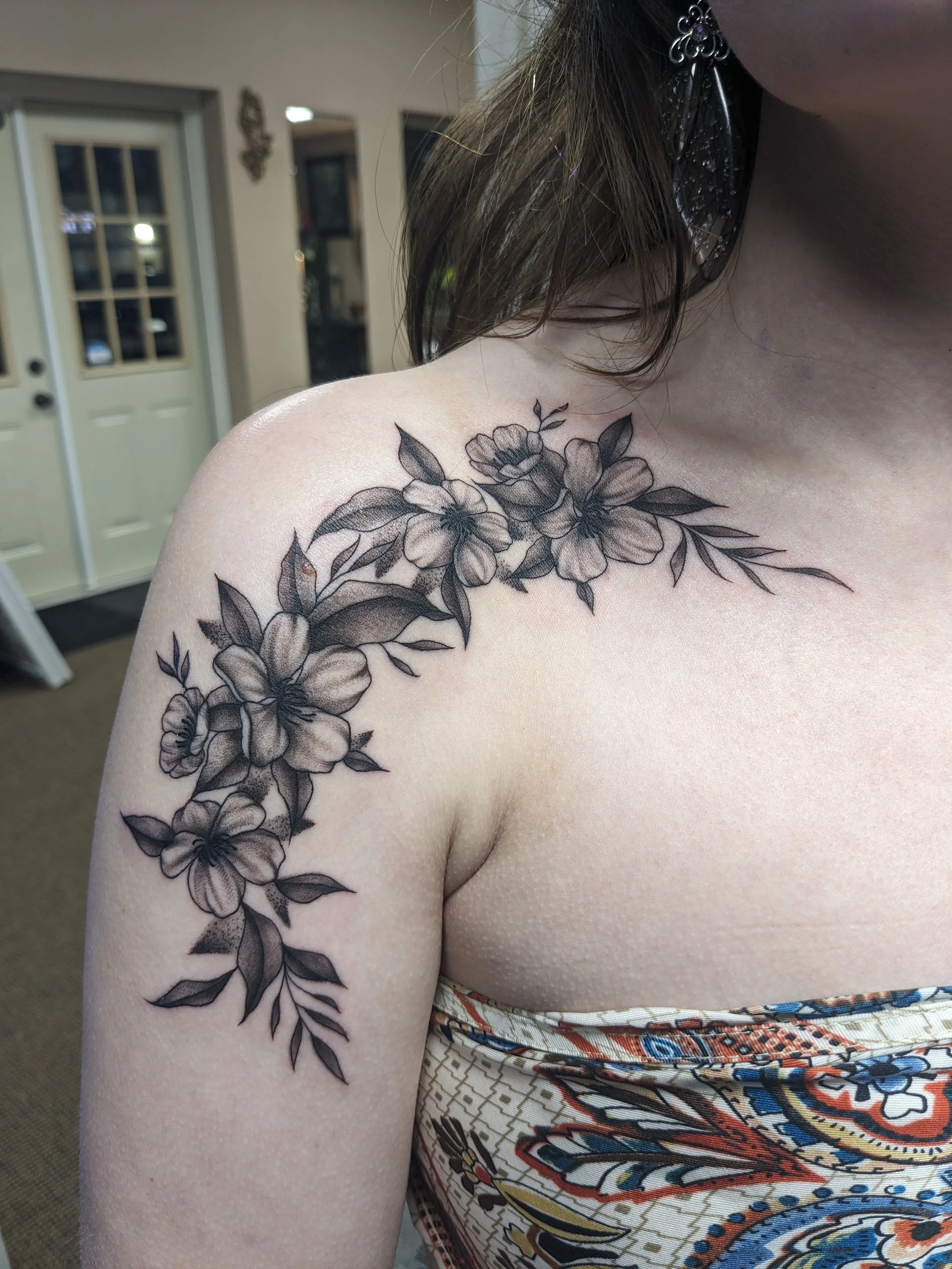 A person's shoulder and upper arm with a black and gray floral tattoo, showing flowers and leaves, next to a patterned fabric with colorful, intricate designs.