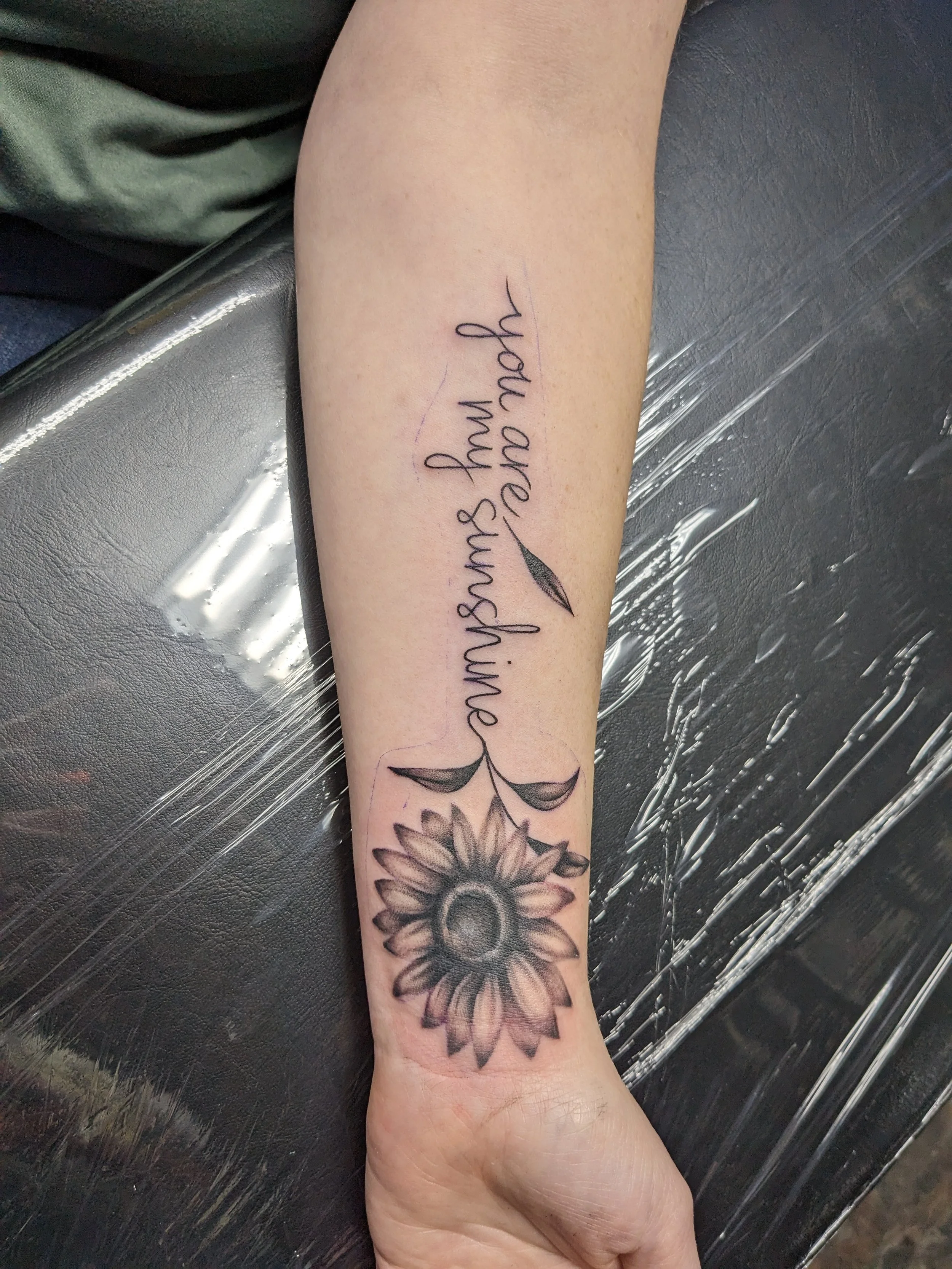 A tattoo on a person's forearm that includes a flower, leaves, and a handwritten-style quote reading 'Banishes and'. The tattoo is in black ink with detailed shading and line work.