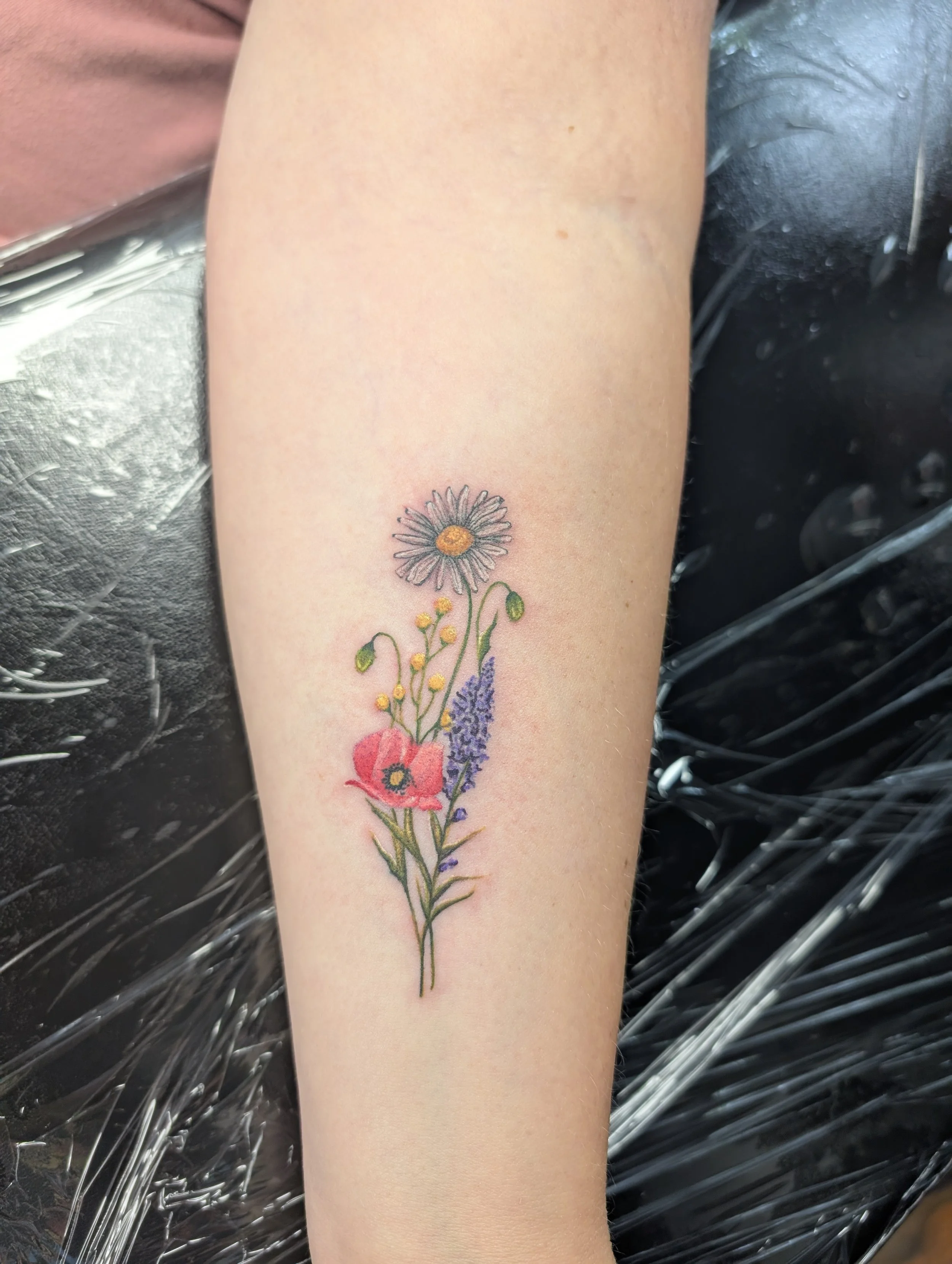 Colorful floral tattoo on person's forearm, featuring daisy, lavender, and other wildflowers, with green stems and leaves.