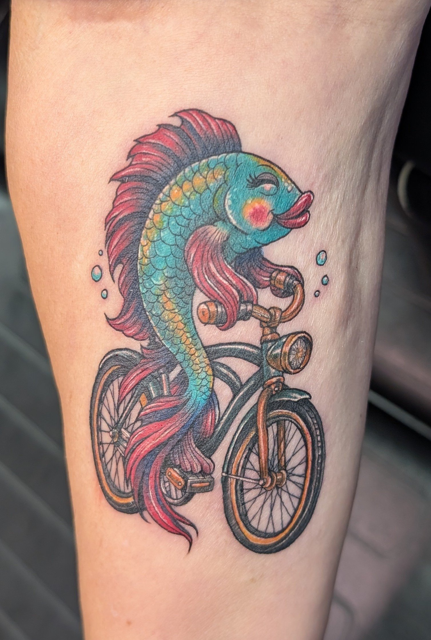 A colorful tattoo of a fish with scales and fins, riding a vintage bicycle, with splashes of water around.