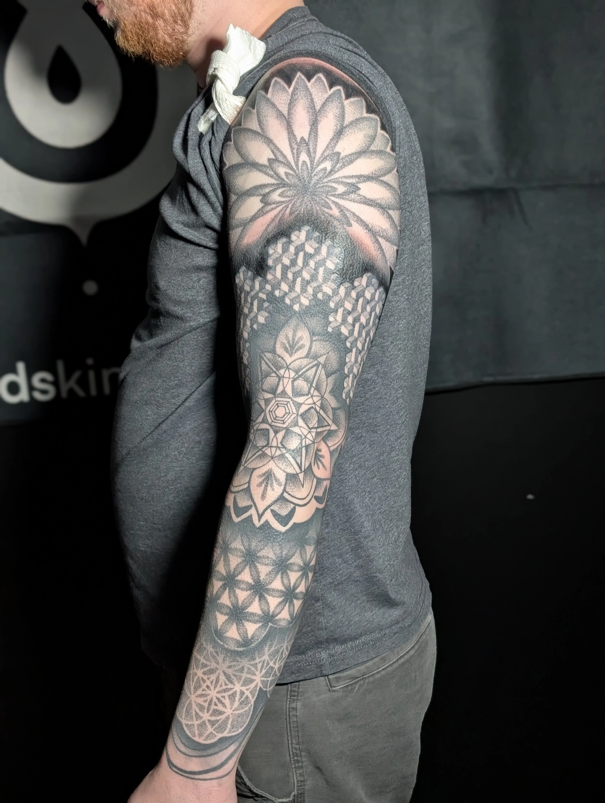 A man with a red beard wearing a gray t-shirt and jeans, showing his arm and shoulder tattoo that features a large floral design with geometric patterns underneath.