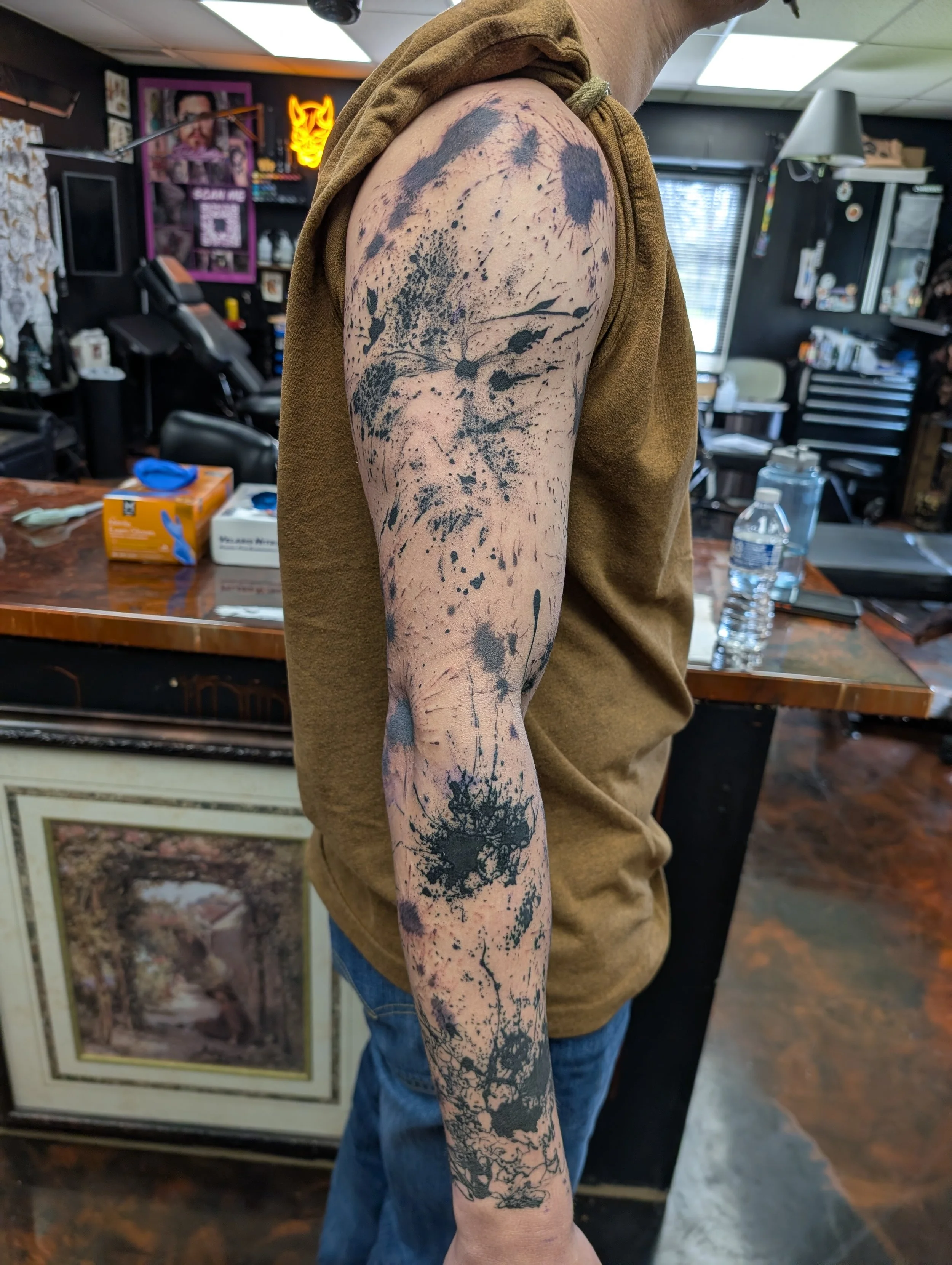 A person's upper arm with a large, black ink splatter tattoo in an abstract style, located in an indoor tattoo studio with tattoos, artwork, and tattoo-related items on the walls.