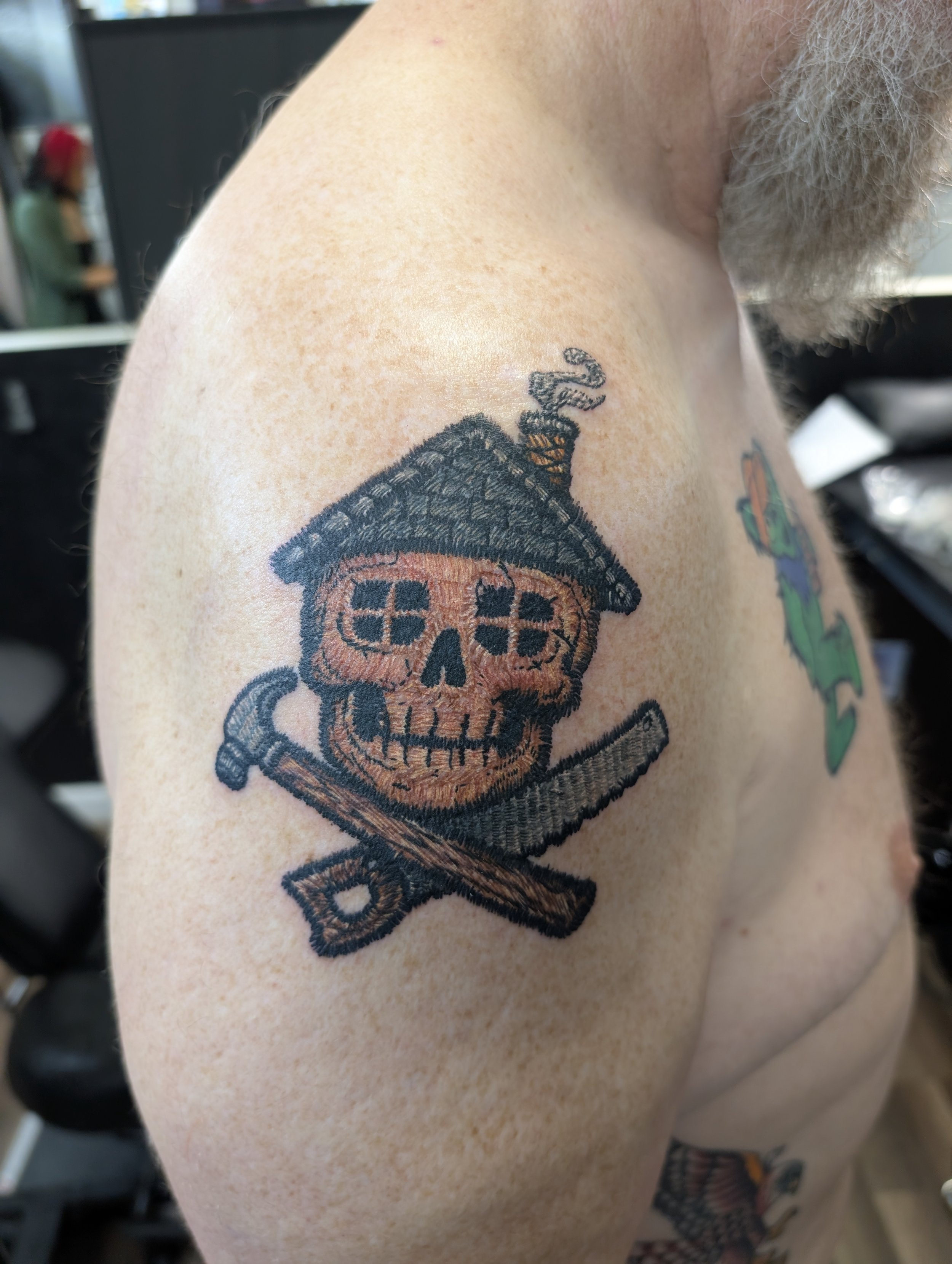 Tattoo of a skull with a house on top and a sword underneath on person's shoulder.