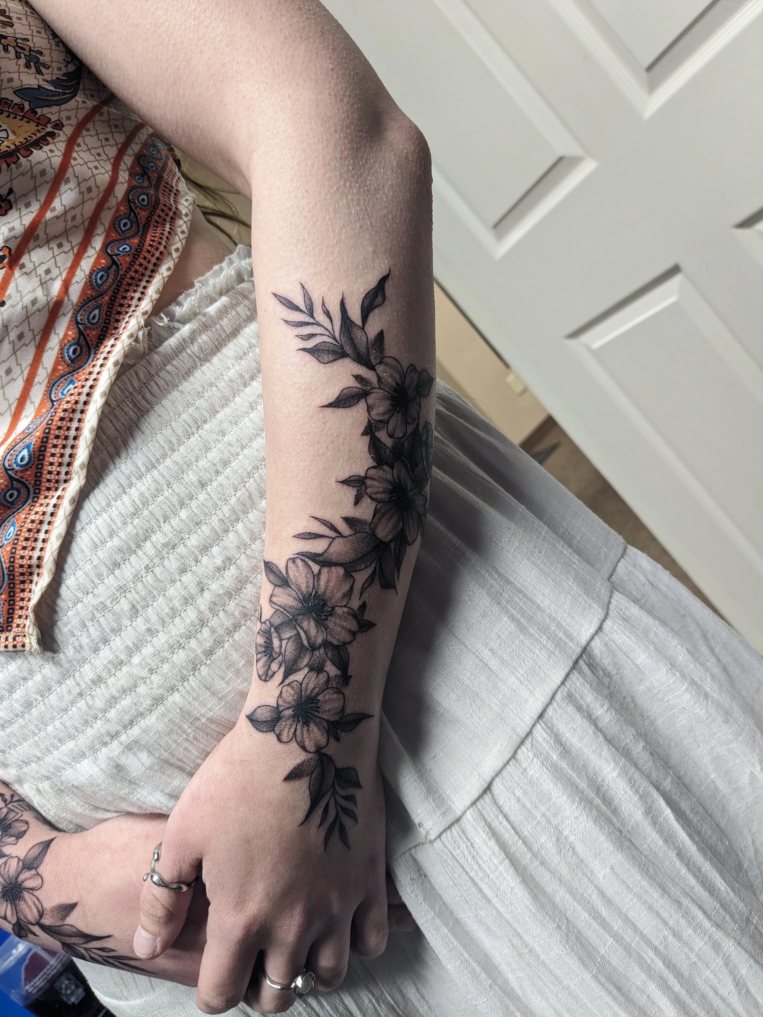 A person's arm and hand with a black floral tattoo covering the forearm and wrist, showing flowers and leaves. The person is wearing rings, a bracelet, and has a patterned shirt and beige pants, sitting on a textured white surface.