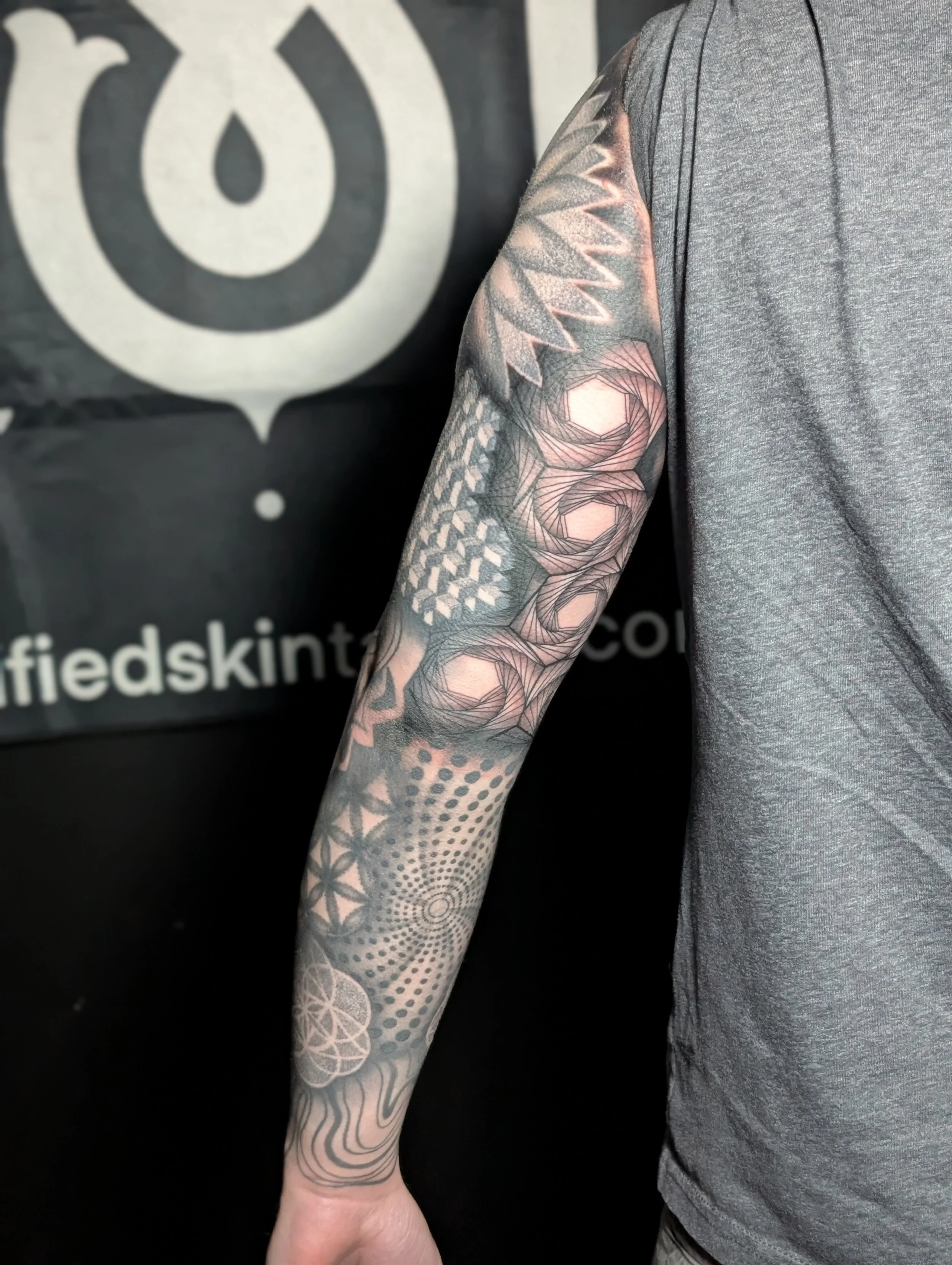 A person with detailed black and pink geometric tattoos on their arm, wearing a gray sleeveless shirt, standing in front of a black background with white text and logo.