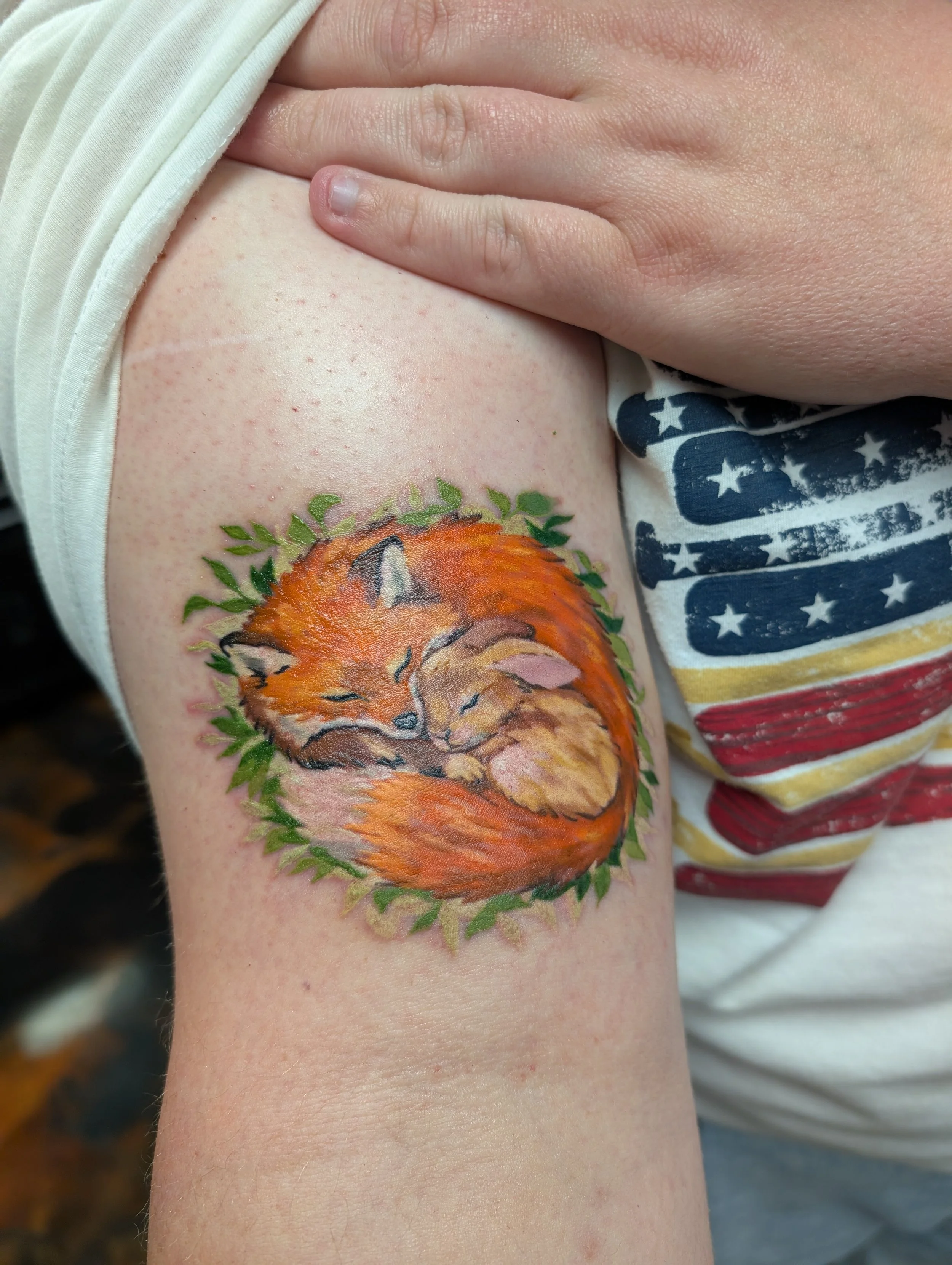 A person's upper arm displaying a colorful fox tattoo surrounded by green leaves, with a hand resting on the shoulder and part of a shirt with an American flag print visible.
