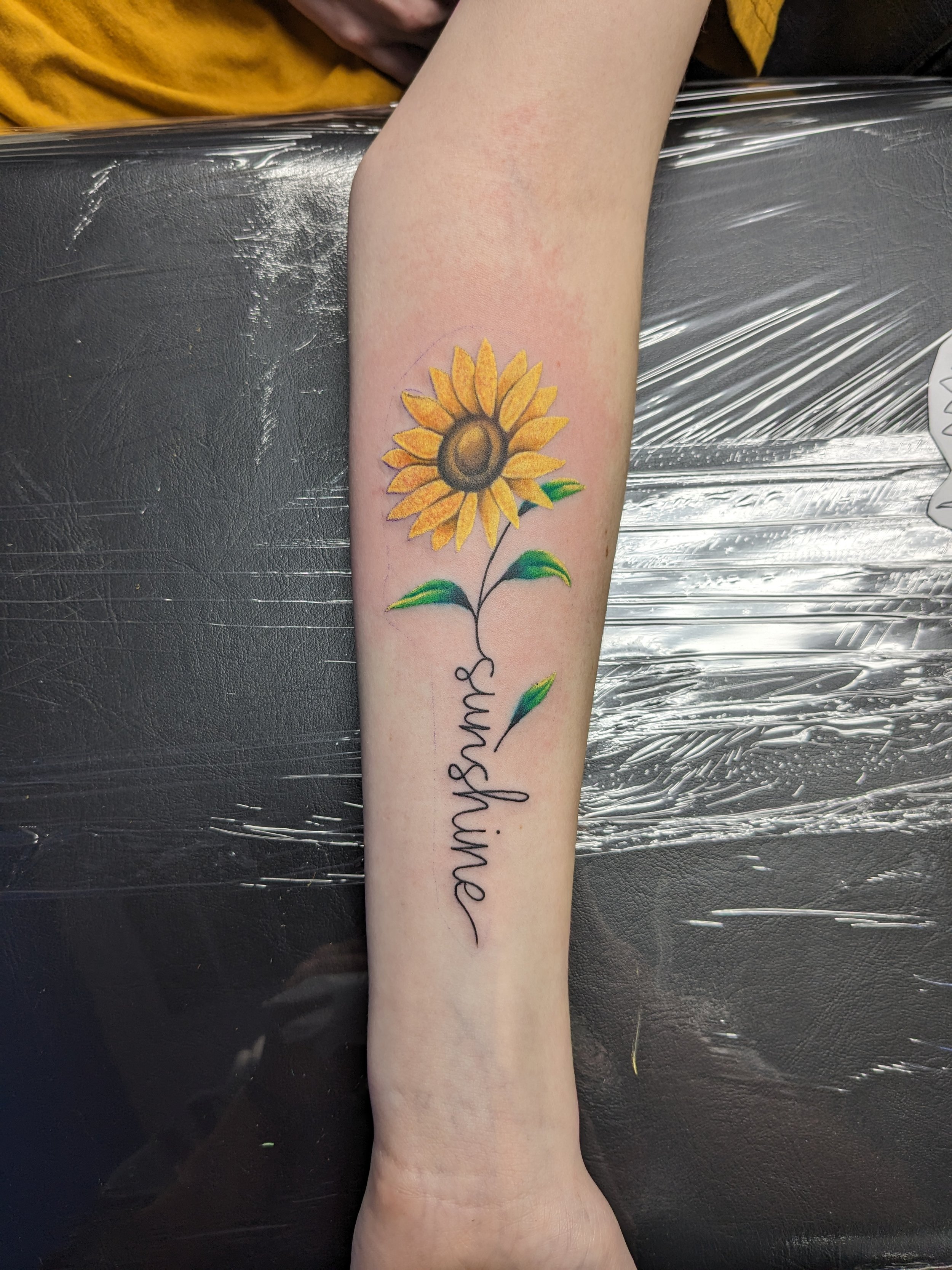 A tattoo of a sunflower with yellow petals, a brown center, and green leaves, along with the word 'sunshine' written in cursive on a person's forearm.