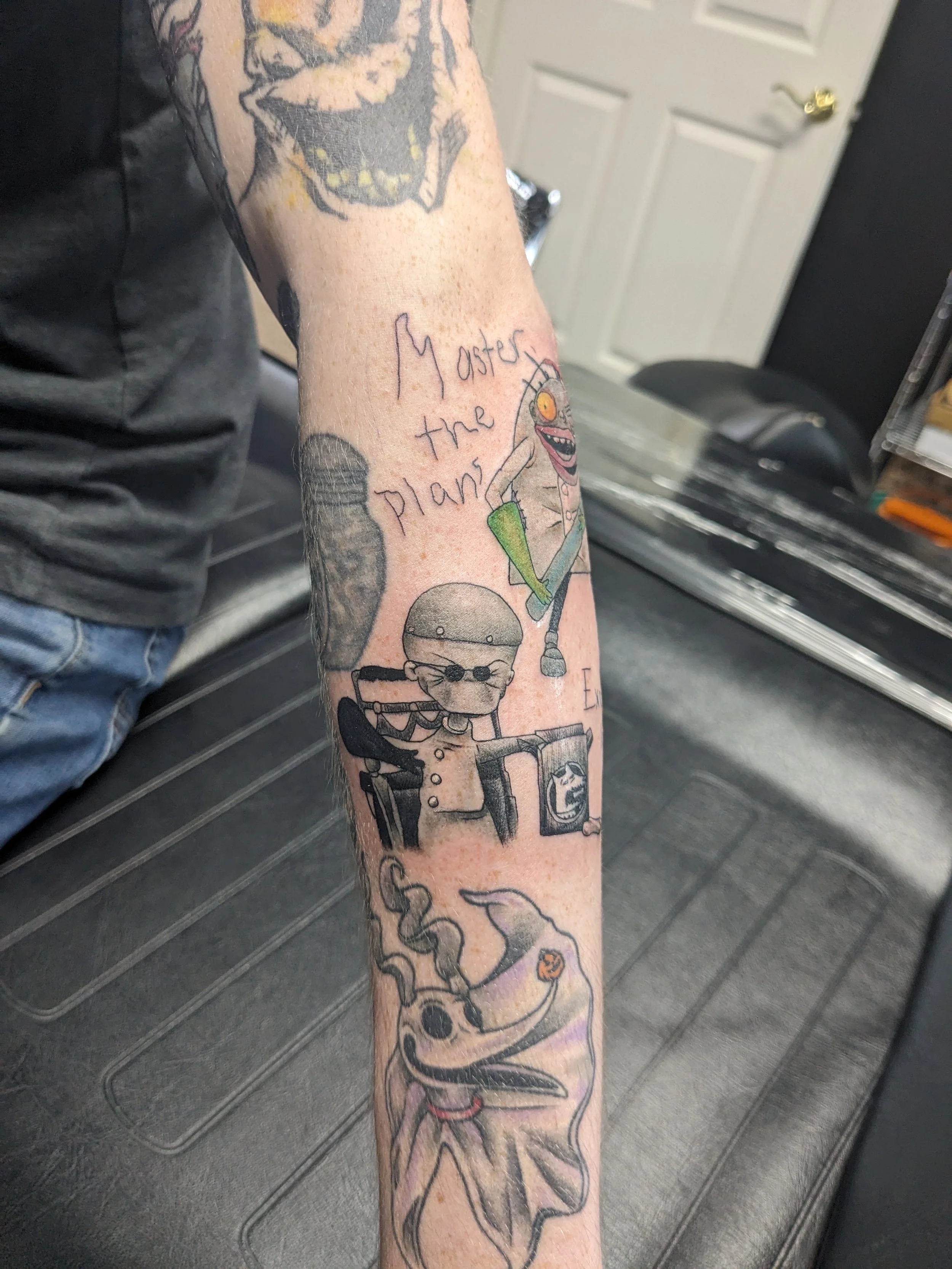 Tattoo on person's arm featuring a cartoonish skeleton character with a big smile, holding a green object, and surrounded by flowing tentacle-like shapes. The tattoo includes the words "Master the plan" written in cursive.