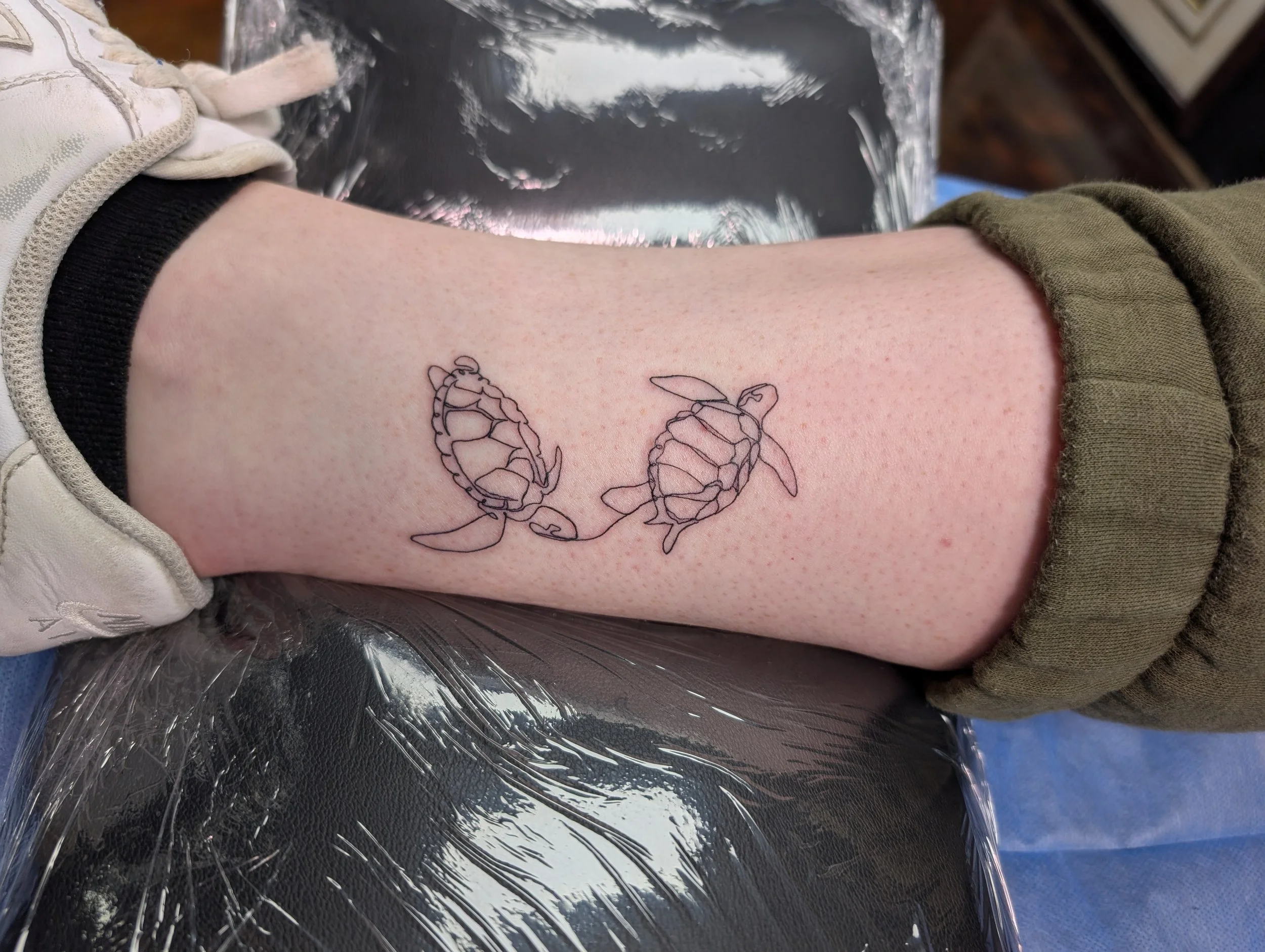 A person's upper arm with a tattoo of two butterflies, a spray of leaves, and a flower, in front of a plastic-covered surface, possibly at a tattoo studio.