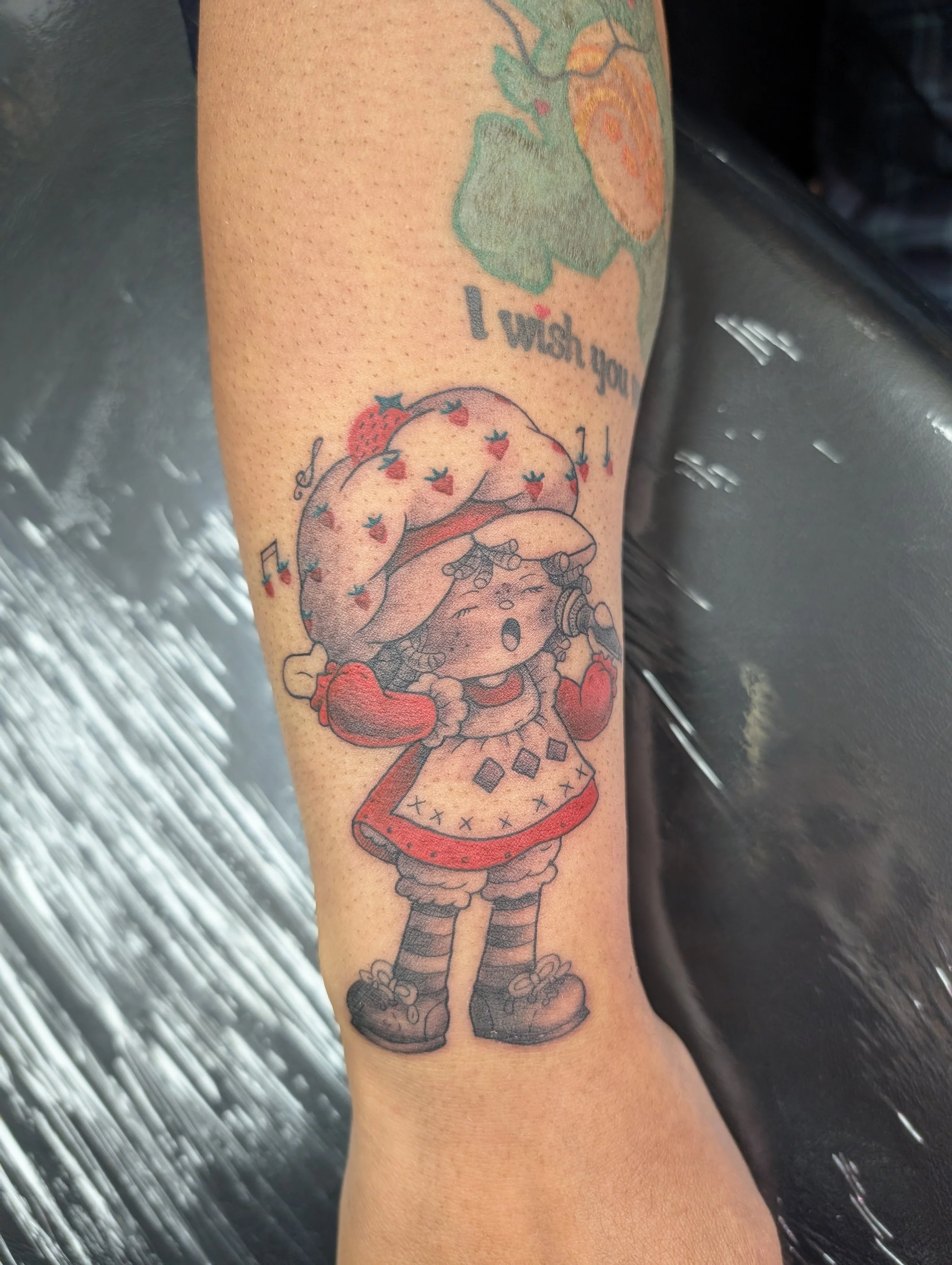 Colorful tattoo of a cartoon girl dressed as Little Red Riding Hood, holding a basket and wearing a hooded cape, on a person's forearm with the text "I wish you" above.