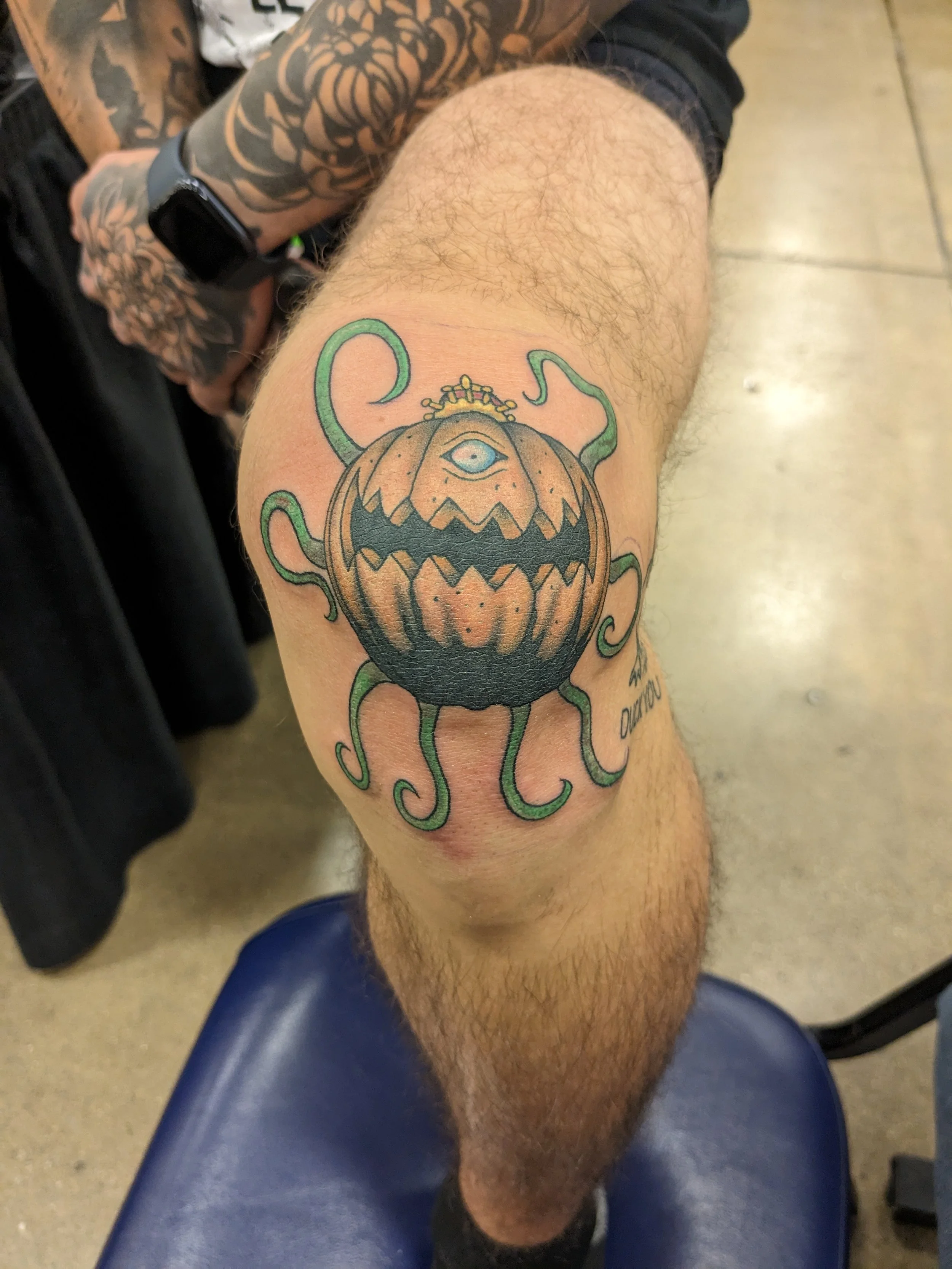 Tattoo of a pumpkin with a third eye and green tentacles on a person's thigh.
