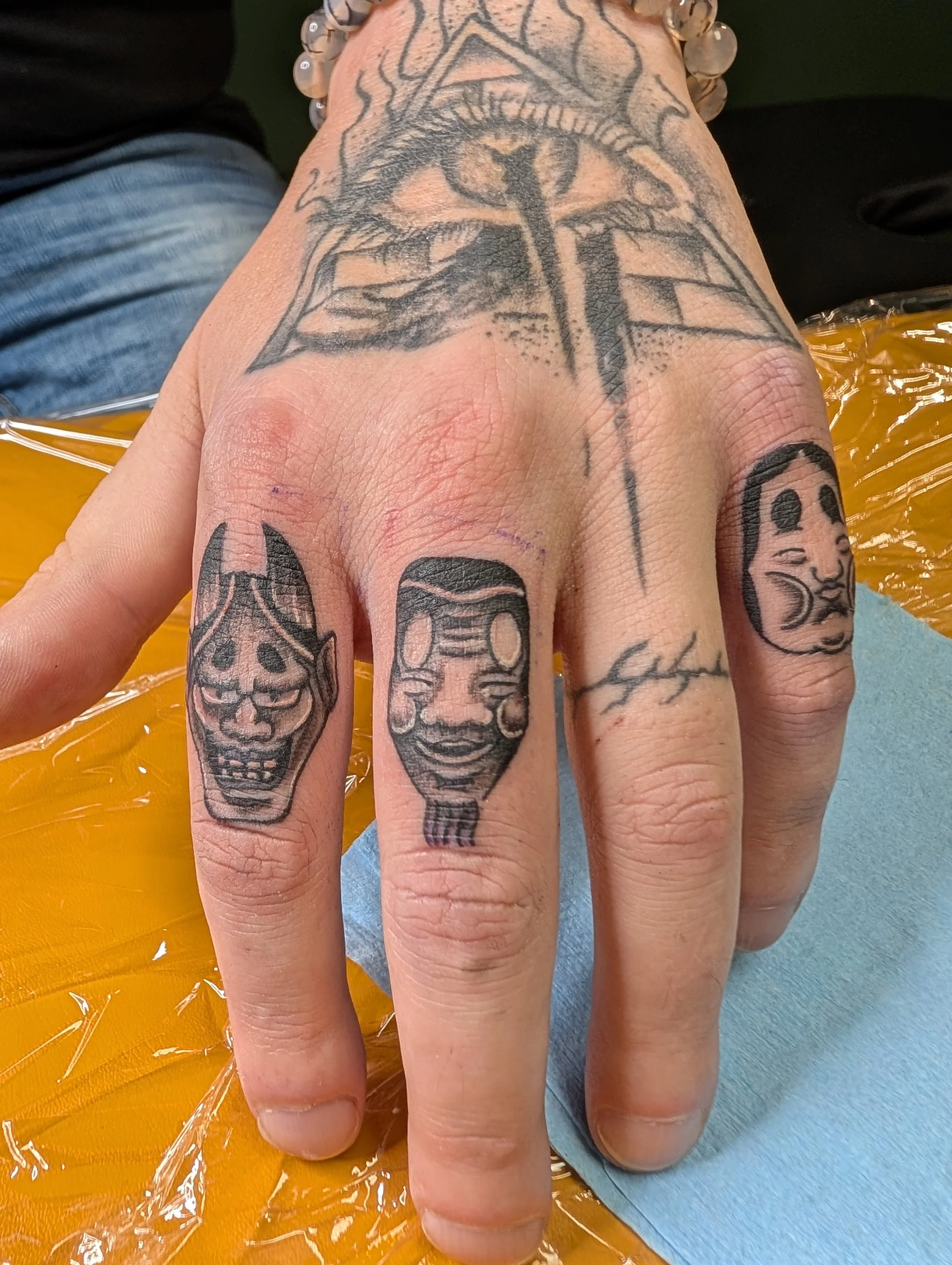 Close-up of a hand with various traditional-style tattoos, including a prominent black and gray dragon on the back of the hand and a smiling face on the finger. The person is wearing a beaded bracelet and is seated on a yellow and orange plastic surf