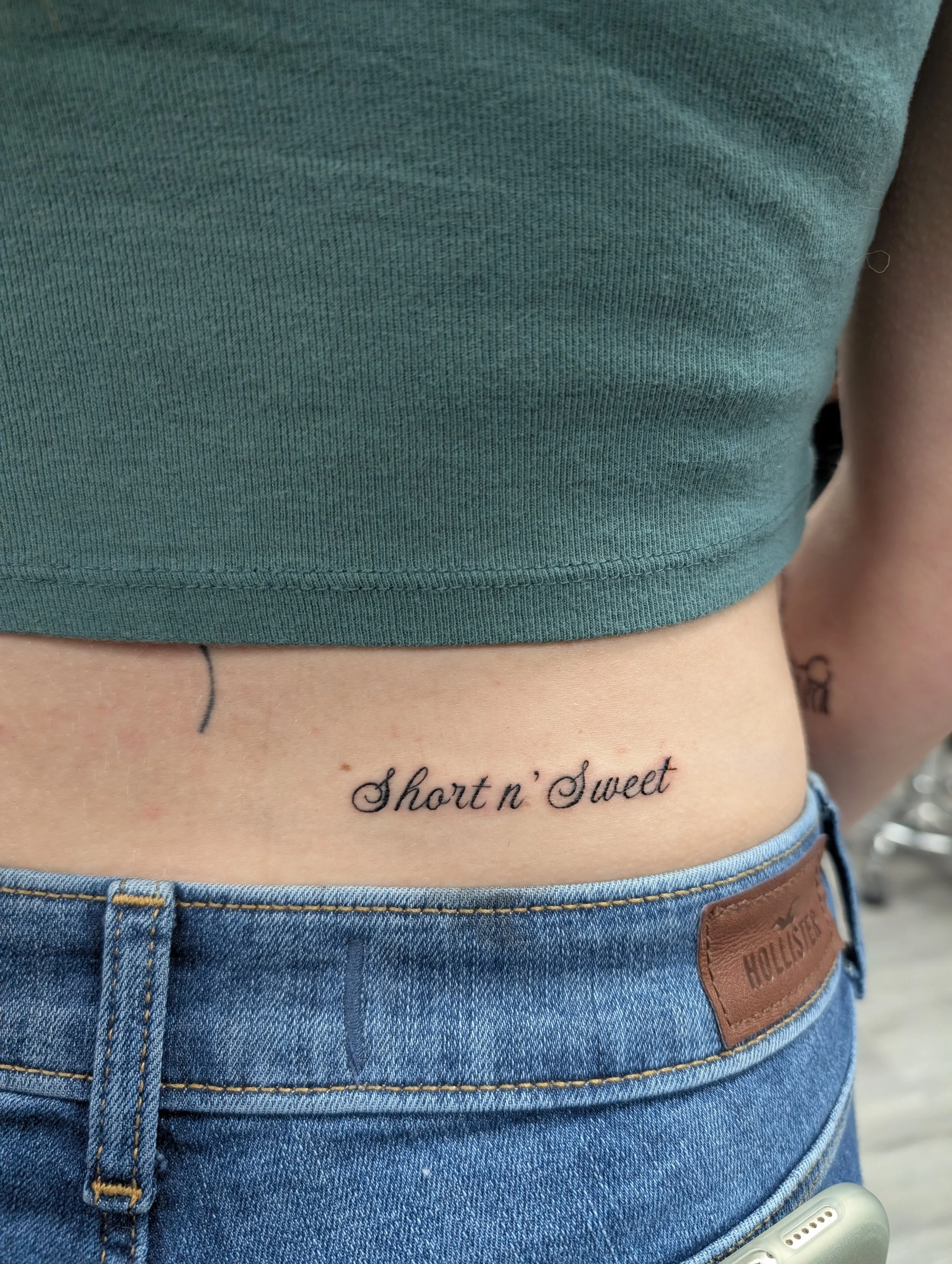 Close-up of a person's midsection wearing a green cropped shirt, blue denim jeans, with a tattoo reading 'Short n' Sweet' on the lower abdomen.