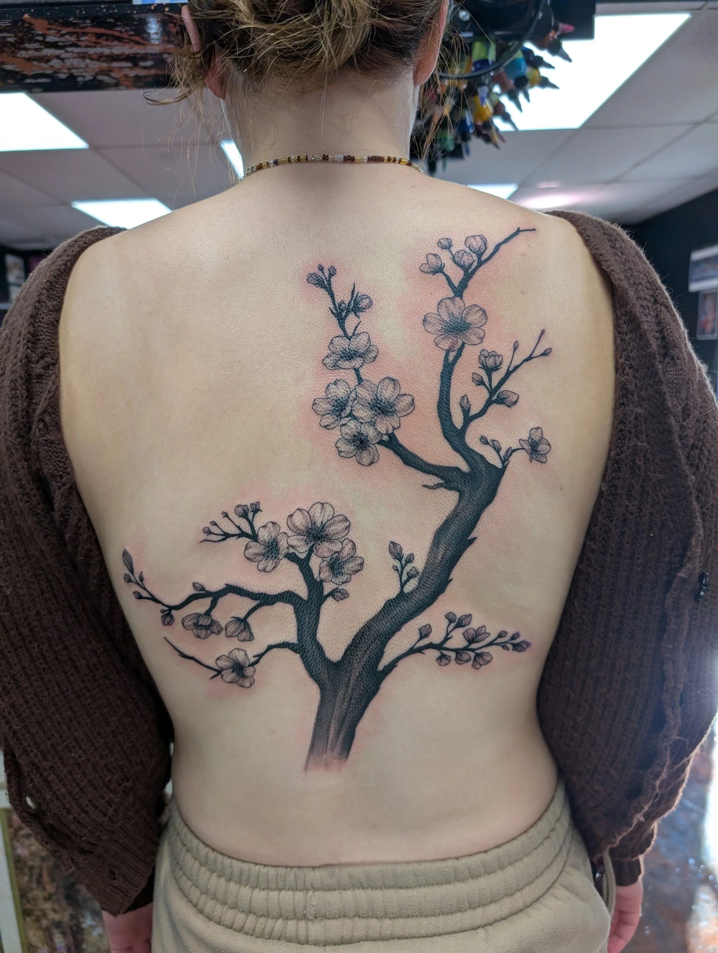 Back view of a woman with a blossom tree tattoo on her back, wearing a brown sweater off her shoulders, a beaded necklace, and beige pants, indoor setting with ceiling lights.